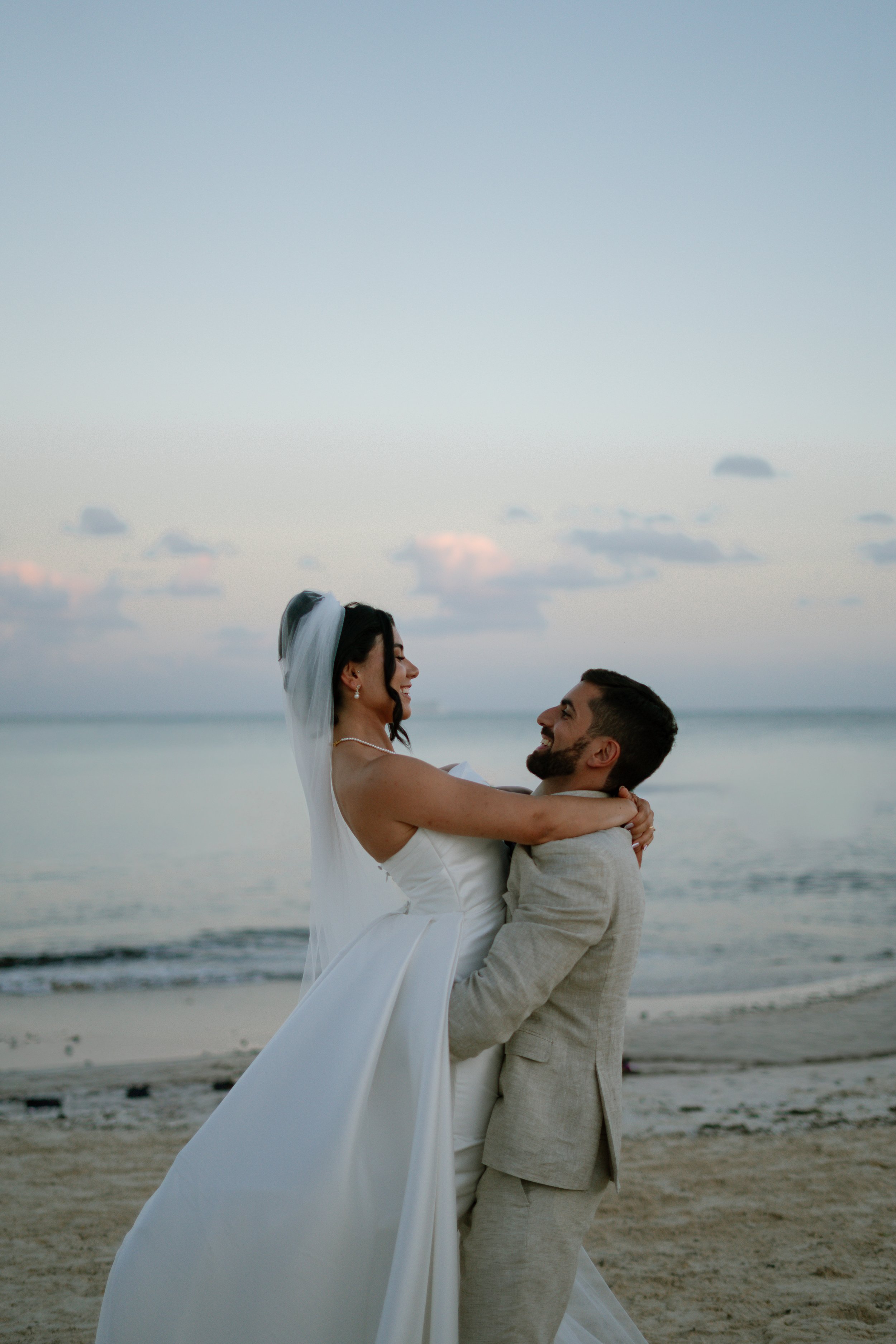 Ein lachendes Brautpaar in Hochzeitskleidung auf einem Strand bei Sonnenuntergang.