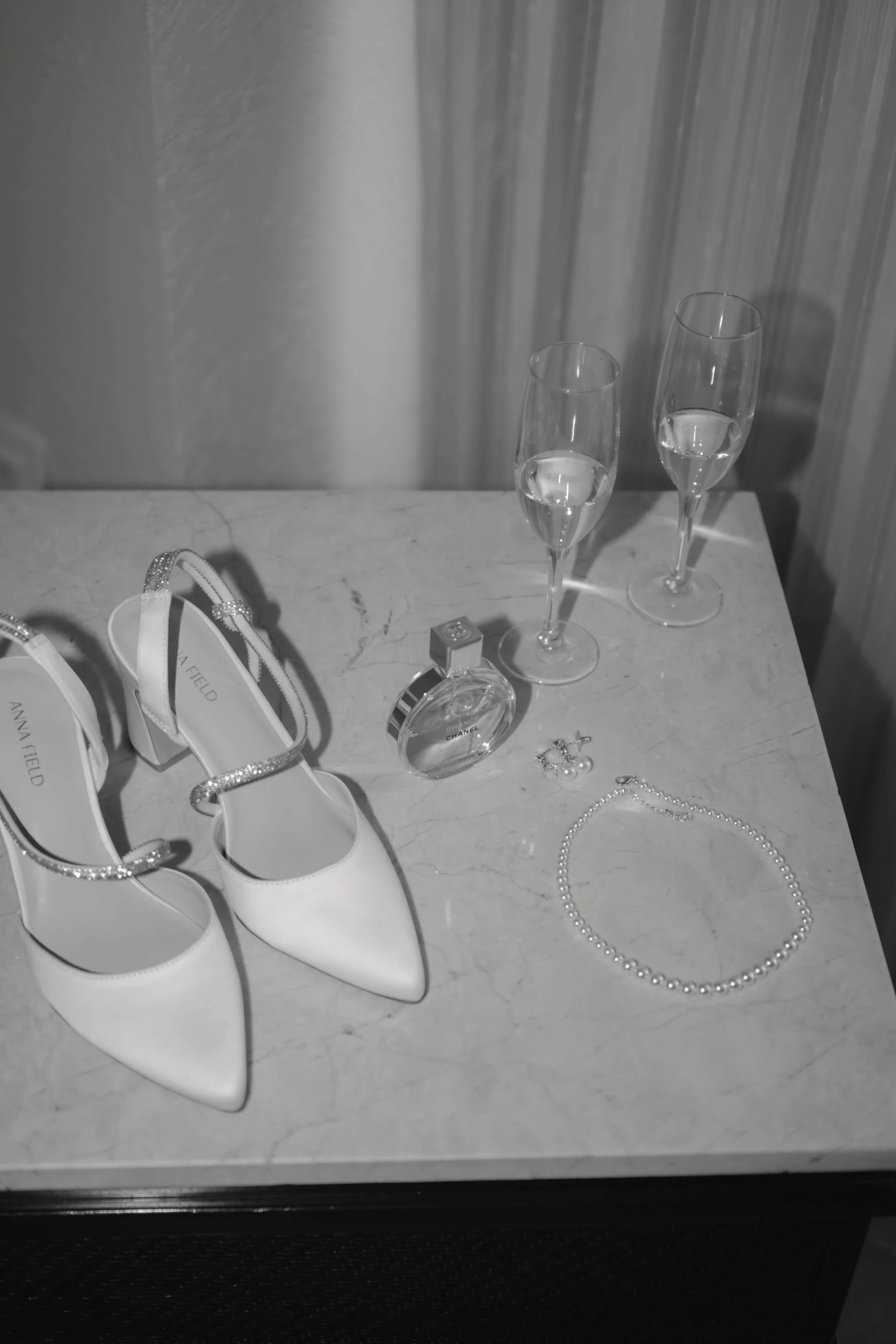 Weiße High Heels, zwei Sektgläser mit Sekt, ein Parfümflakon, ein Paar Ohrringe und eine Perlenkette auf einer weißen Marmorplatte