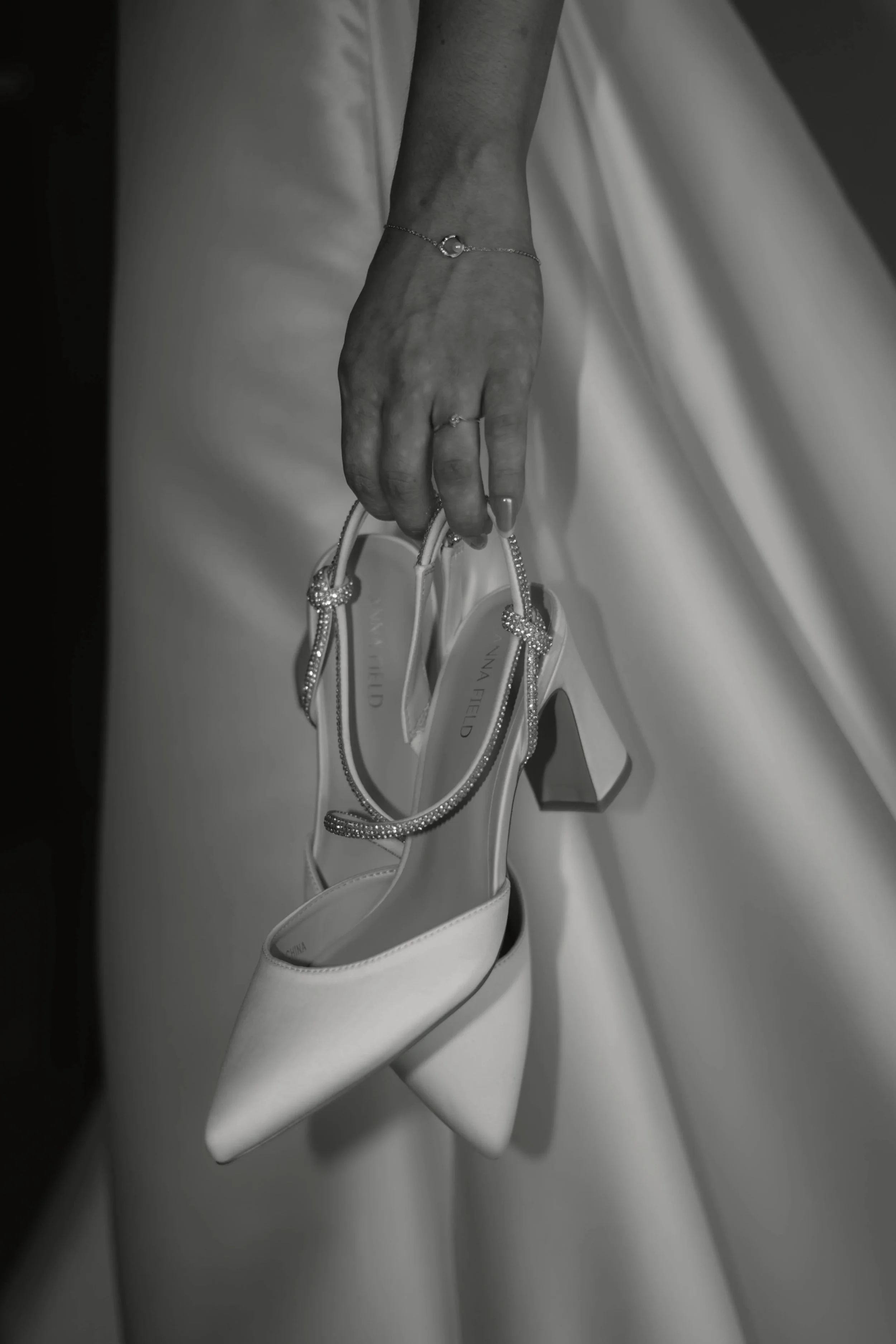 Ein Hand hält ein Paar weiße High Heels mit spitzen Zehen, bei einem eleganten Anlass. Die Hand trägt eine Schmuckkette und ein Armband.