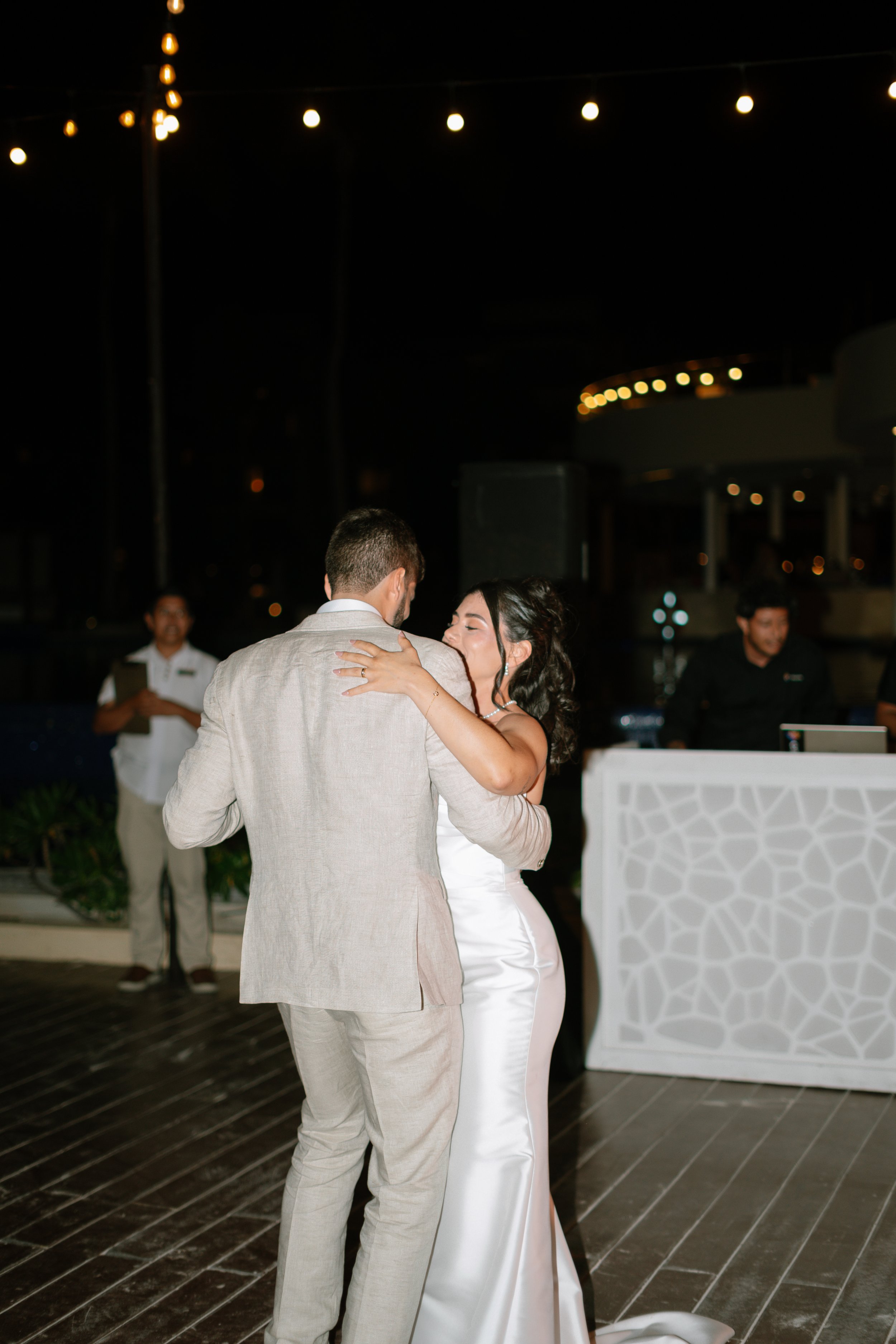 Paare tanzen bei einer Hochzeitsfeier im Freien bei Nacht, mit Lichterketten im Hintergrund.