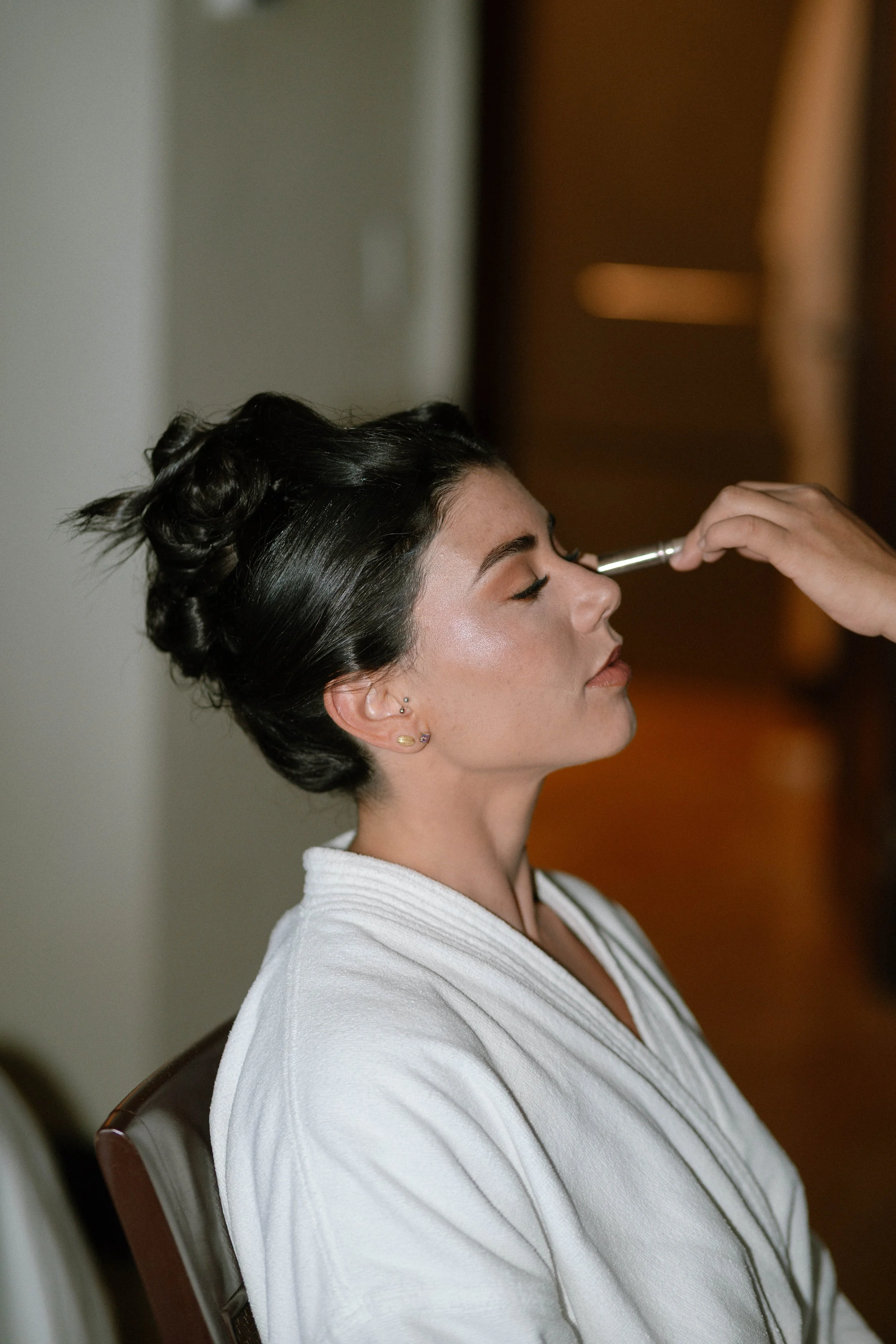 Frau mit geschminktem Gesicht sitzt mit geschlossenen Augen, während ein Makeup-Künstler einen Pinsel auf ihre Nase tupft.