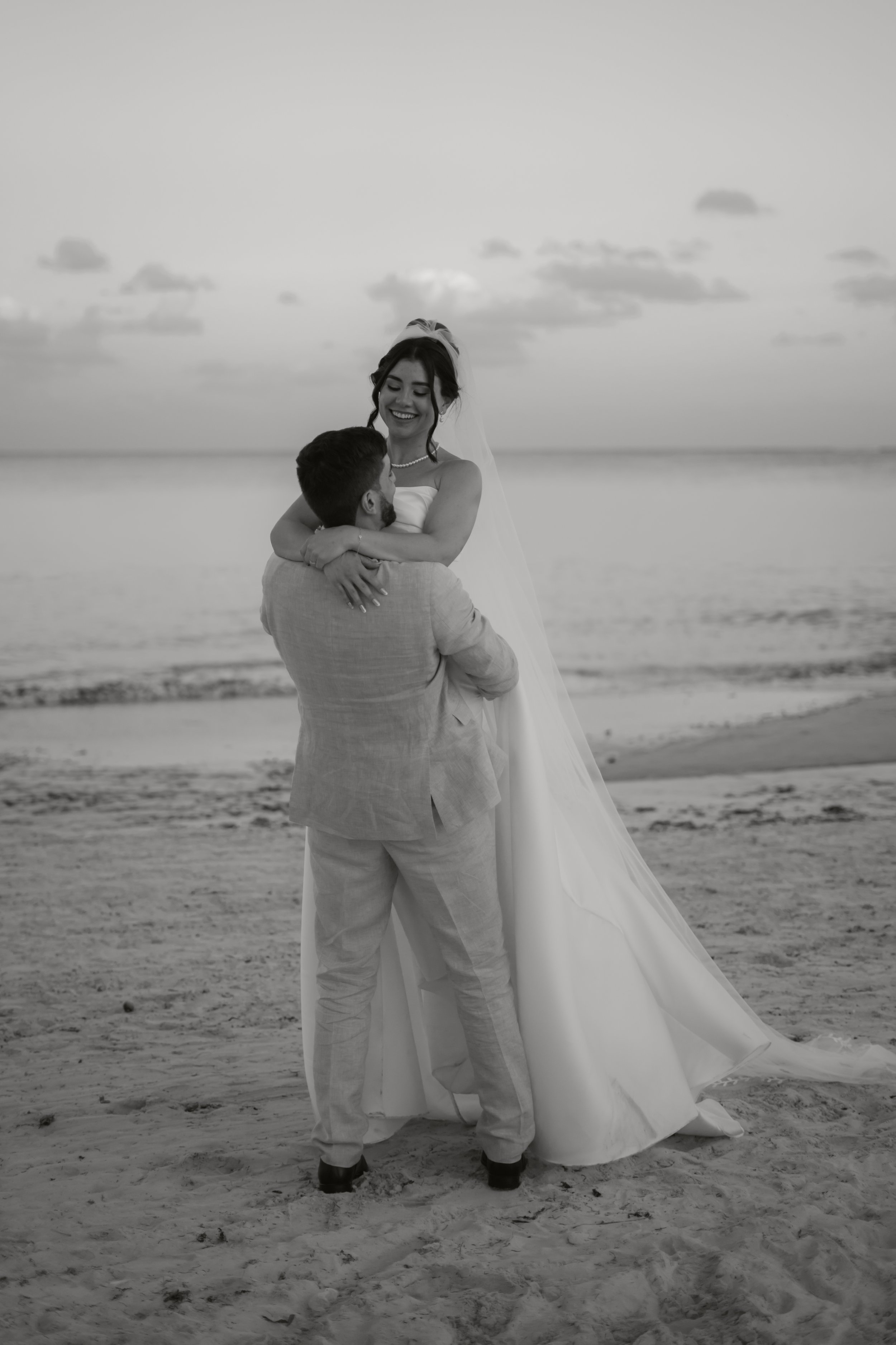 Ein glückliches Brautpaar am Strand, schwarz-weiß fotografiert, mit der Braut in einem weißen Kleid, die umarmt wird.
