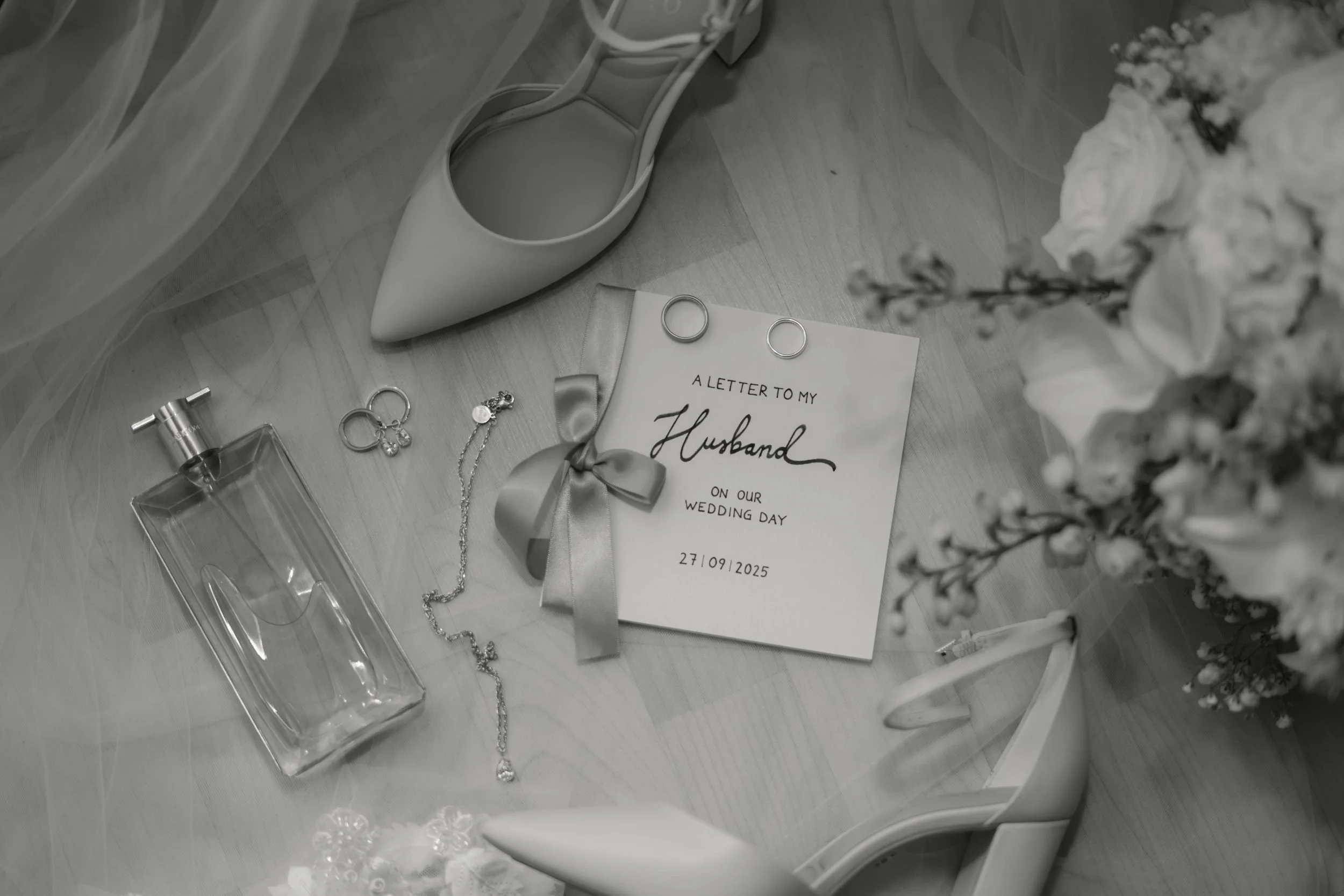 Schwarz-weiß-Bild zeigt Brautschmuck, eine Karte mit dem Text „A letter to my husband on our wedding day 27/09/2025“, ein Paar weiße Schuhe, eine Glückwunschkarte, einen Blumenstrauß, und andere Hochzeitsaccessoires auf einem Holztisch.