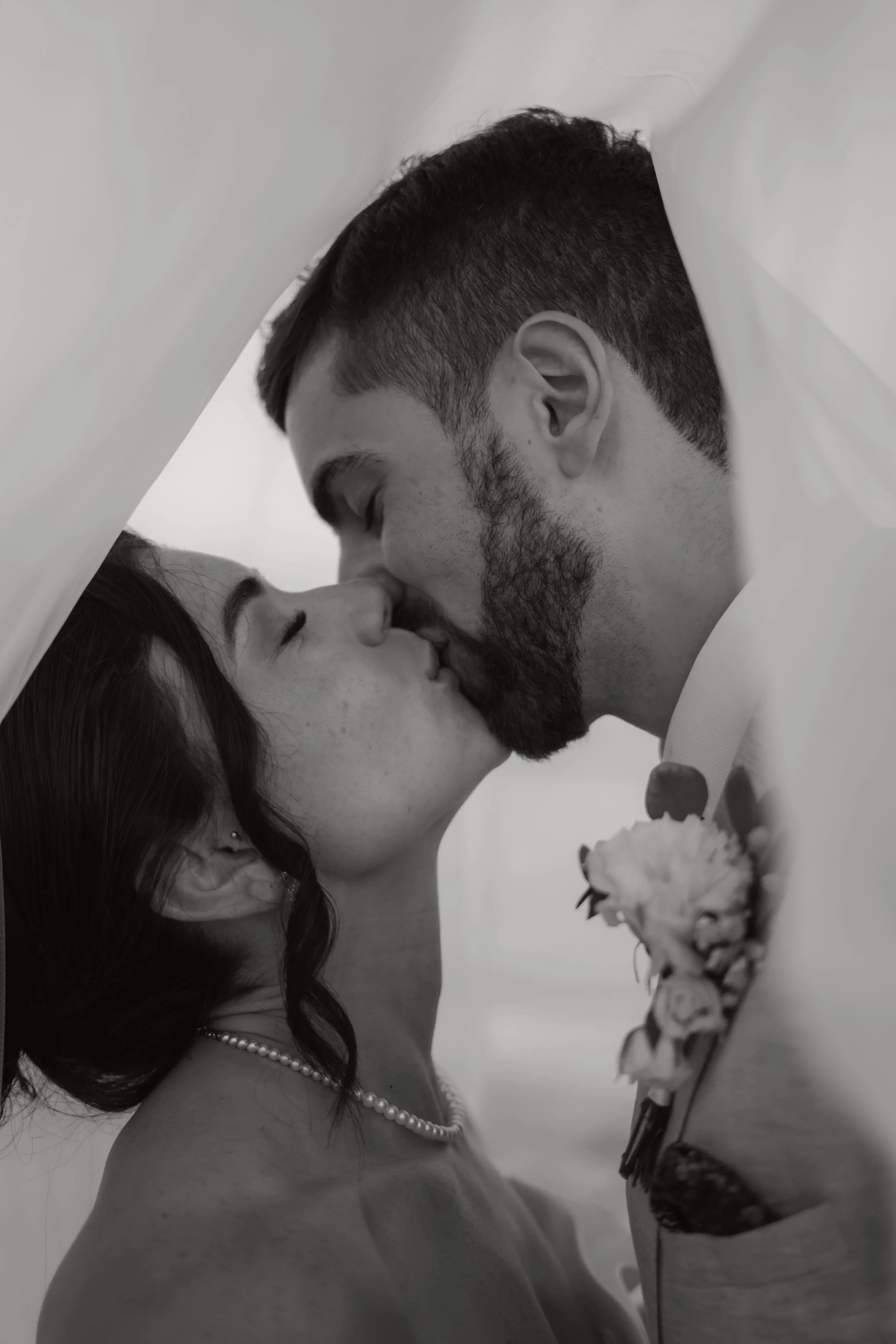 Ein Brautpaar bei einer Hochzeitszeremonie, sie küssen sich unter einem weißen Tuch. Er trägt einen Anzug mit einer Rose als Boutonnière, sie trägt eine Perlenkette und hat dunkle, lockige Haare.