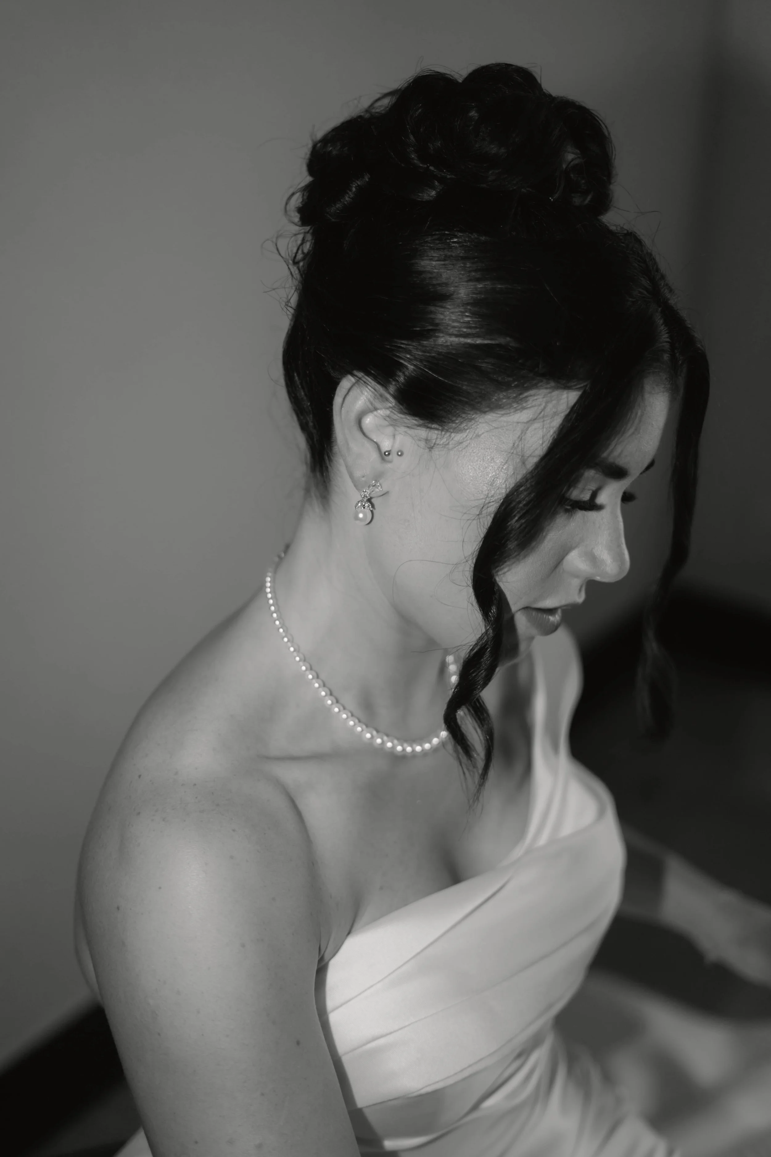 Schwarz-weiß-Fotografie einer Frau in einem weißen Kleid, die nach unten schaut, mit dunklem, hochgestecktem Haar, Perlenschmuck und mehreren Ohrpiercings.