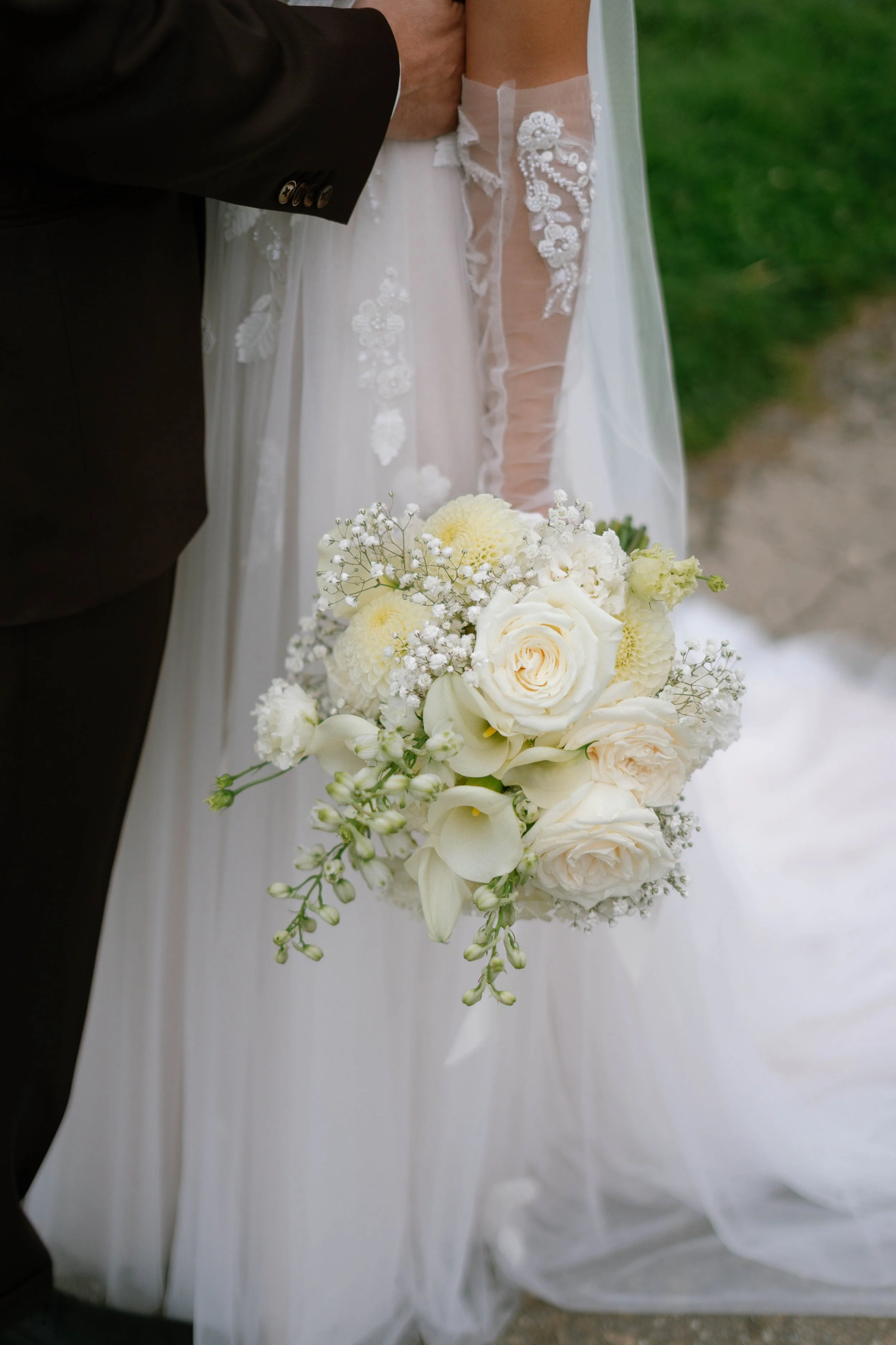Nahaufnahme eines Brautstraußes mit weißen Blumen, gehalten von einer Braut in einem weißen Hochzeitskleid. Im Hintergrund sind die Beine eines Bräutigams in einem schwarzen Anzug sichtbar.