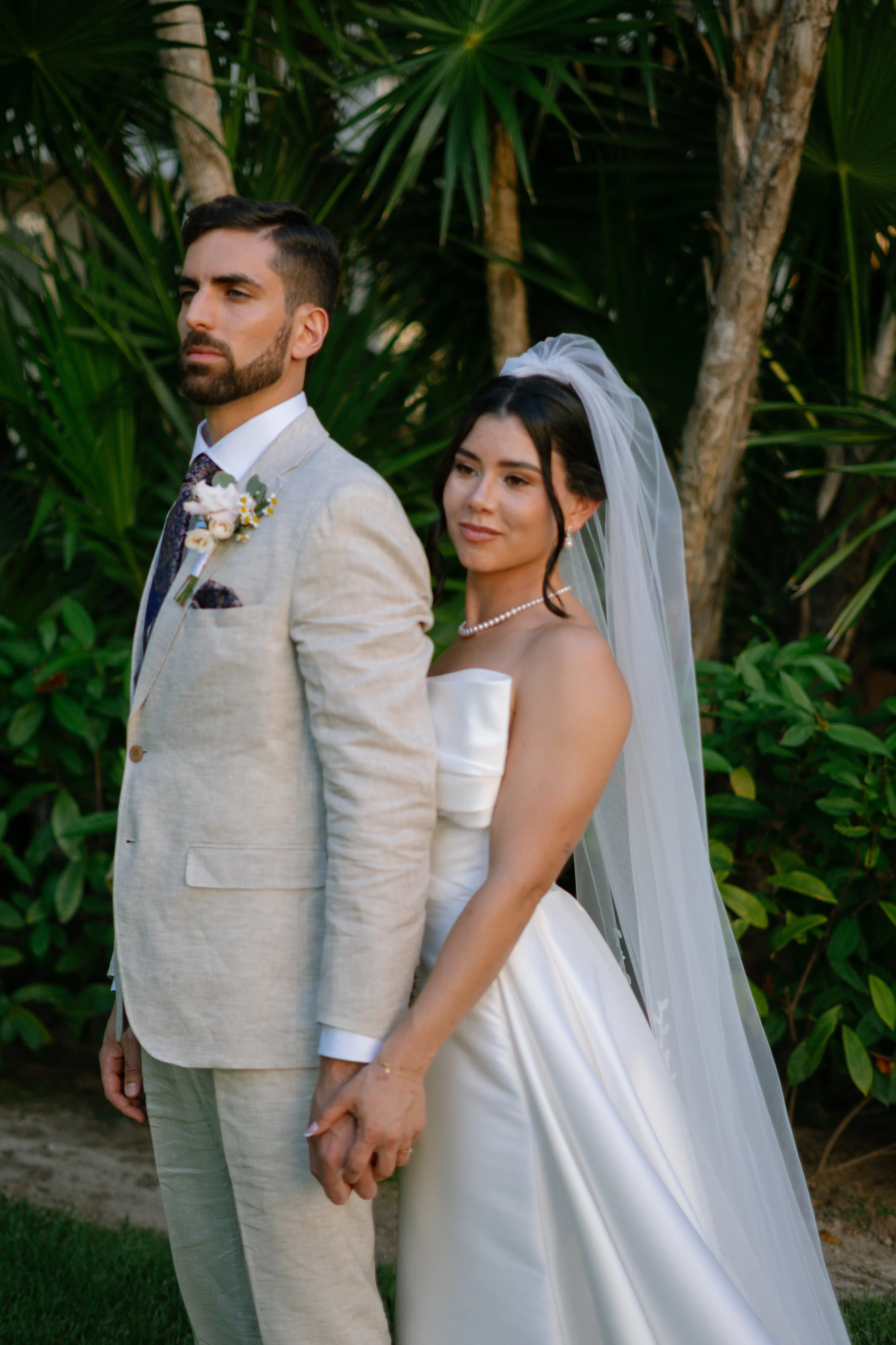 Ein Brautpaar in formeller Kleidung, die Hände haltend, im Freien vor grünen Pflanzen und Bäumen, im Hintergrund.