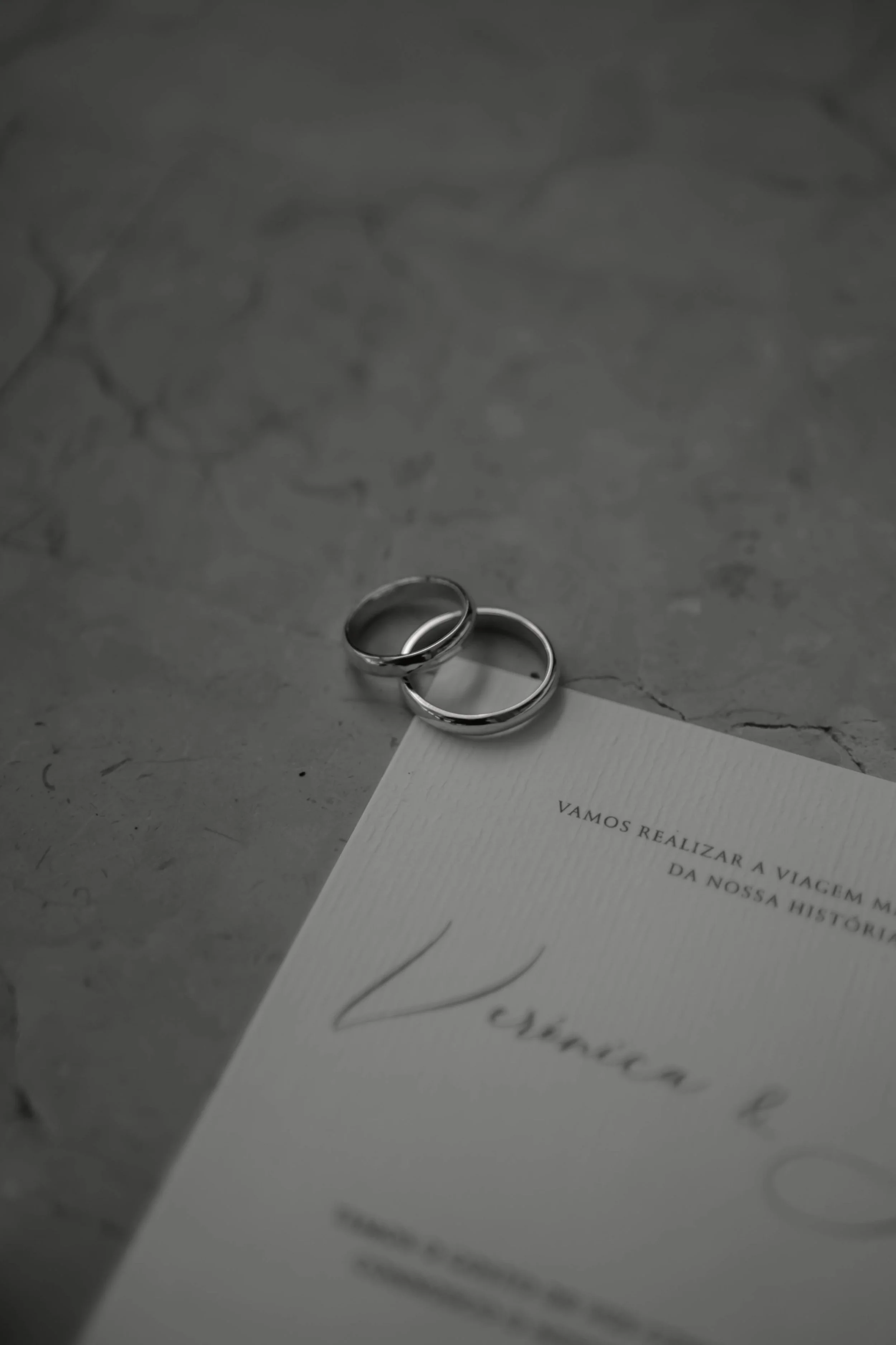 Zwei Eheringe liegen auf einem Papier mit portugiesischem Text auf einer grauen, marmorierten Oberfläche.