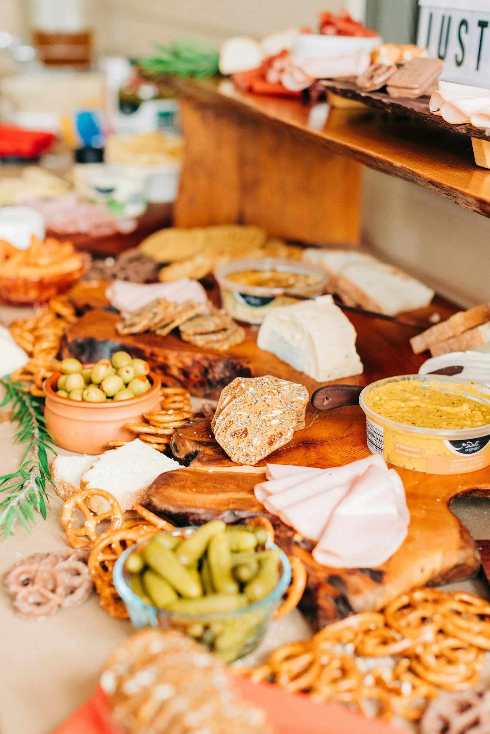 A variety of cheeses, crackers, olives, pretzels, hummus, sliced meats, and other snacks arranged on a wooden table for a party or gathering.