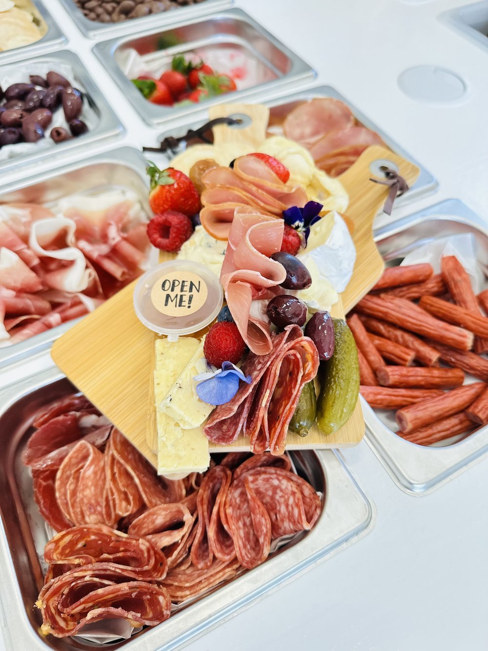 A platter of assorted meats, cheeses, and fruits garnished with edible flowers on a wooden board, surrounded by containers of olives, strawberries, cherry tomatoes, and cured meats.