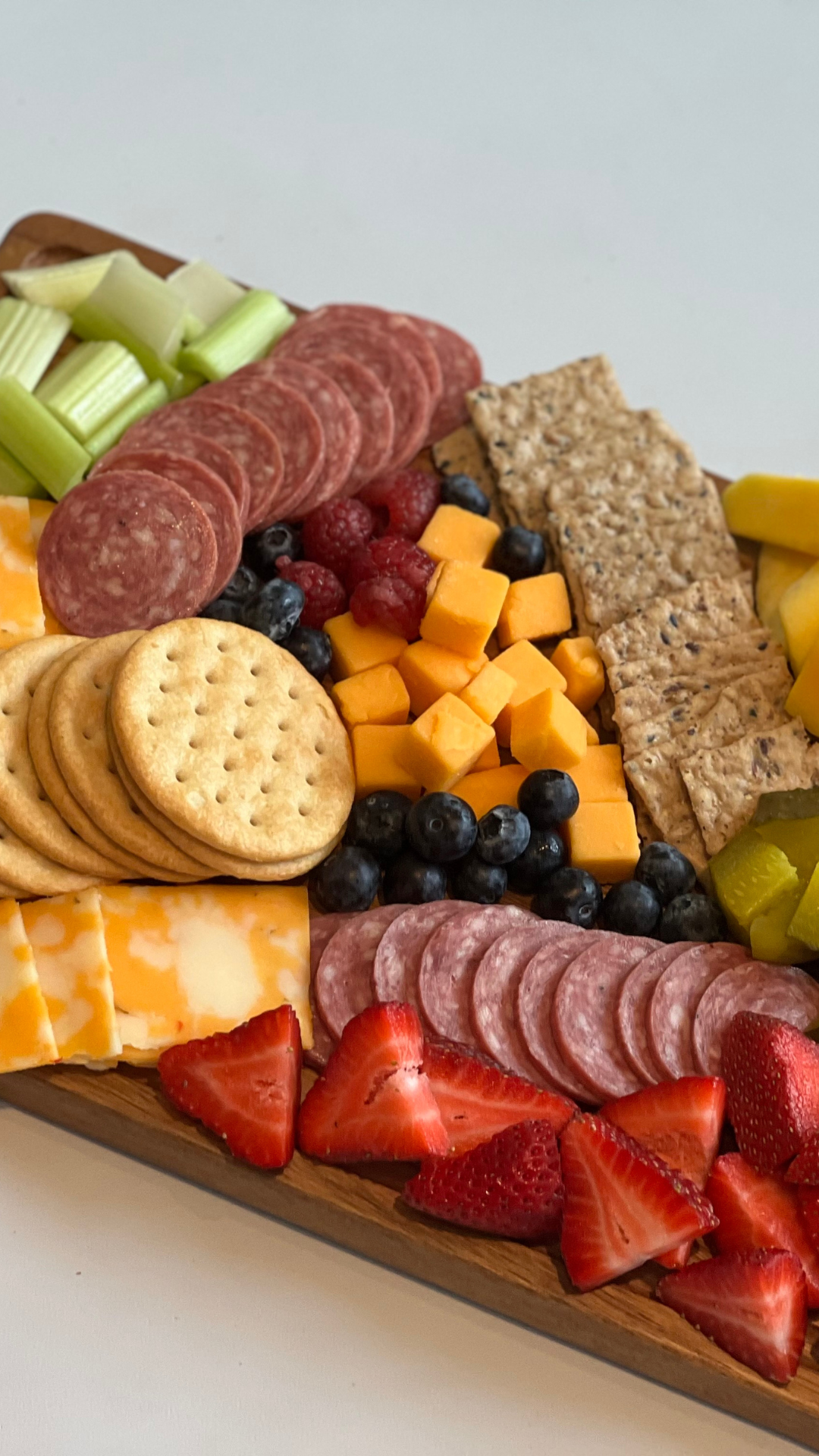 Cheese, strawberries, blueberries, raspberries, crackers, sliced salami, sliced cheese, cubed cheddar cheese, granola bars, celery sticks, pineapple chunks, and other assorted fruit and snack items arranged on a tray.