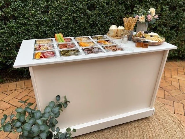 A white outdoor buffet table with various dishes, vegetables, condiments, bread, and accompaniments set against a green hedge backdrop.
