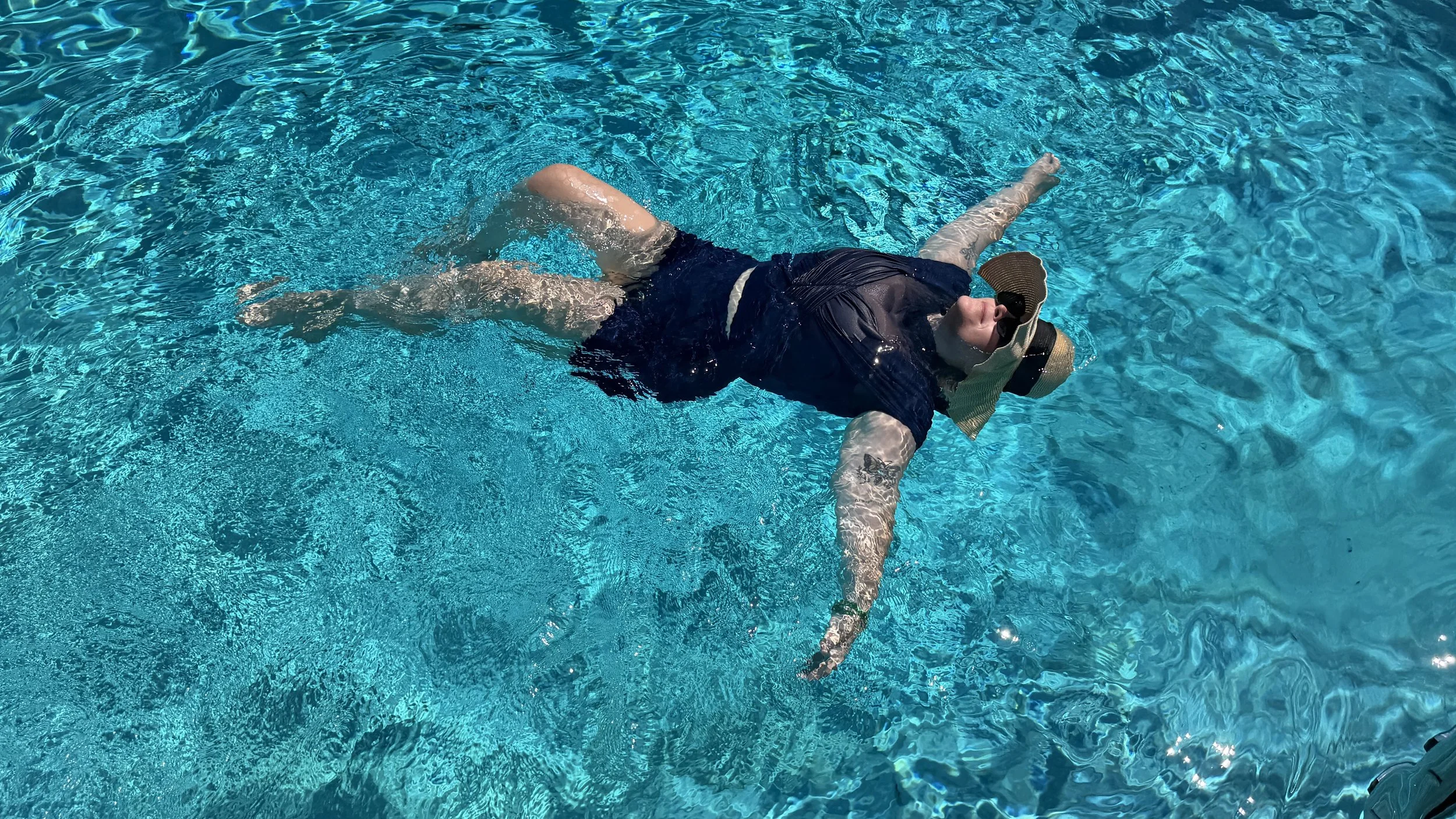 A woman floating on her back in a swimming pool, wearing a full coverage navy swim top, navy swim shorts, a navy sun hat, and sunglasses.