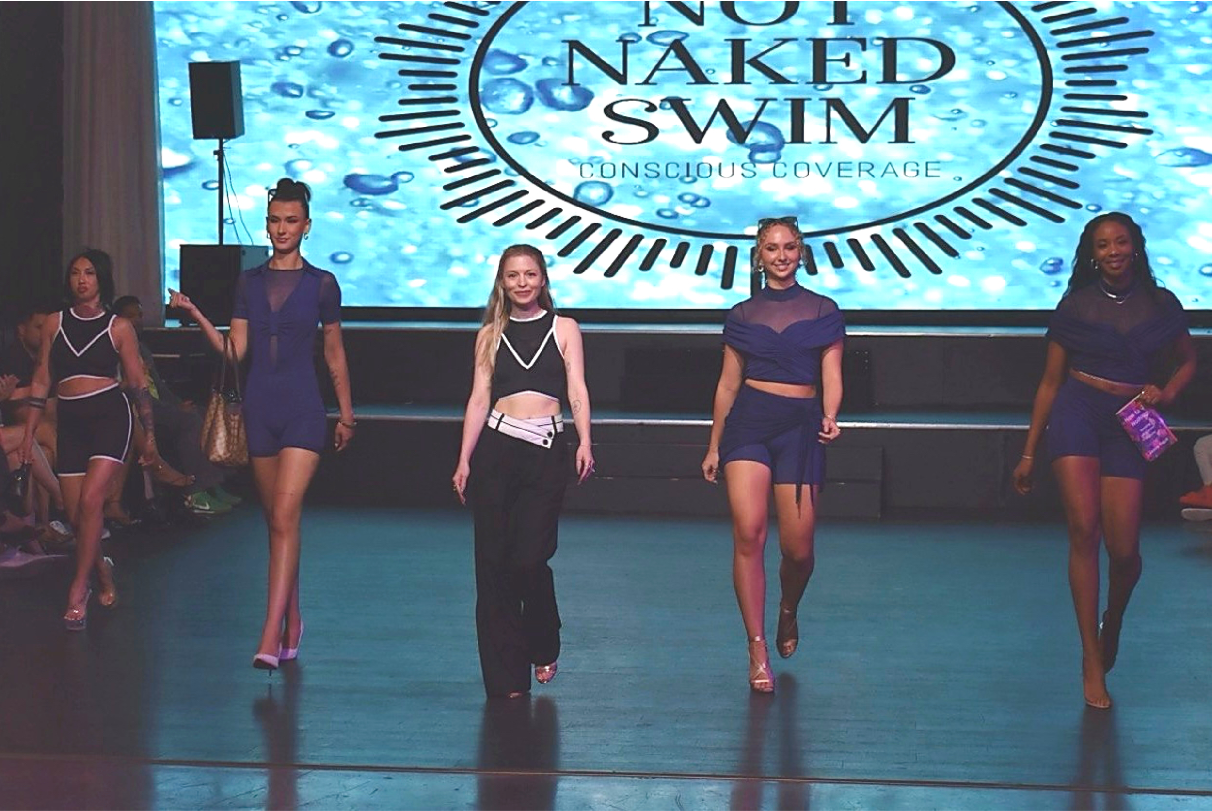 A group of five women walking on a runway during a fashion show, with a large screen behind them displaying the text 'Not Naked Swim' and water bubbles background.