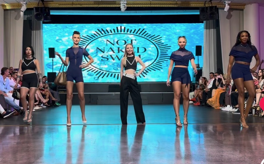 Models walking on runway during a fashion show, with audience on both sides and a large digital screen displaying 'Not Naked Swims' in the background.