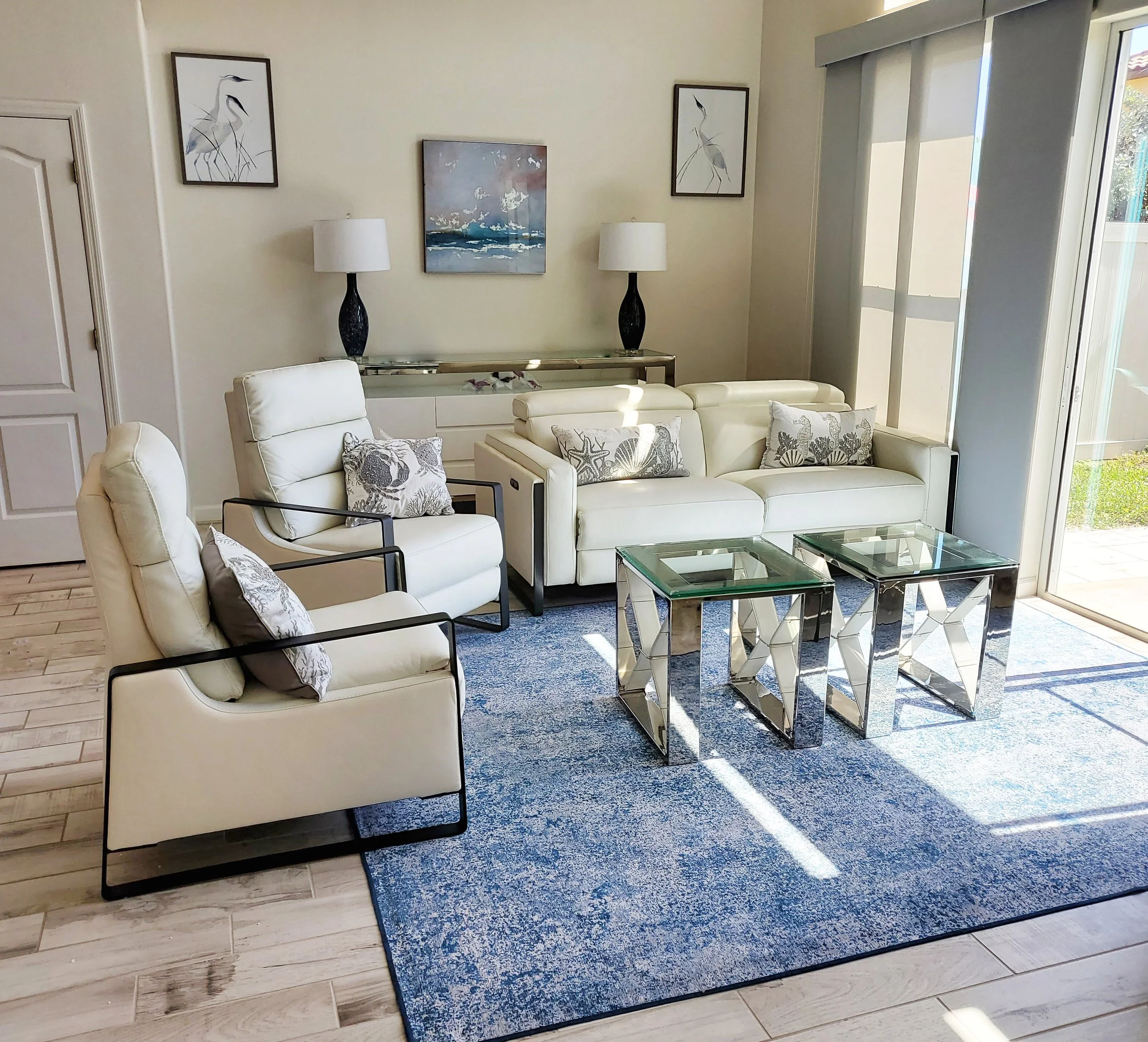 A modern living room with white furniture, decorative pillows, glass-top tables, artwork on the wall, and large sliding glass doors letting in natural light.