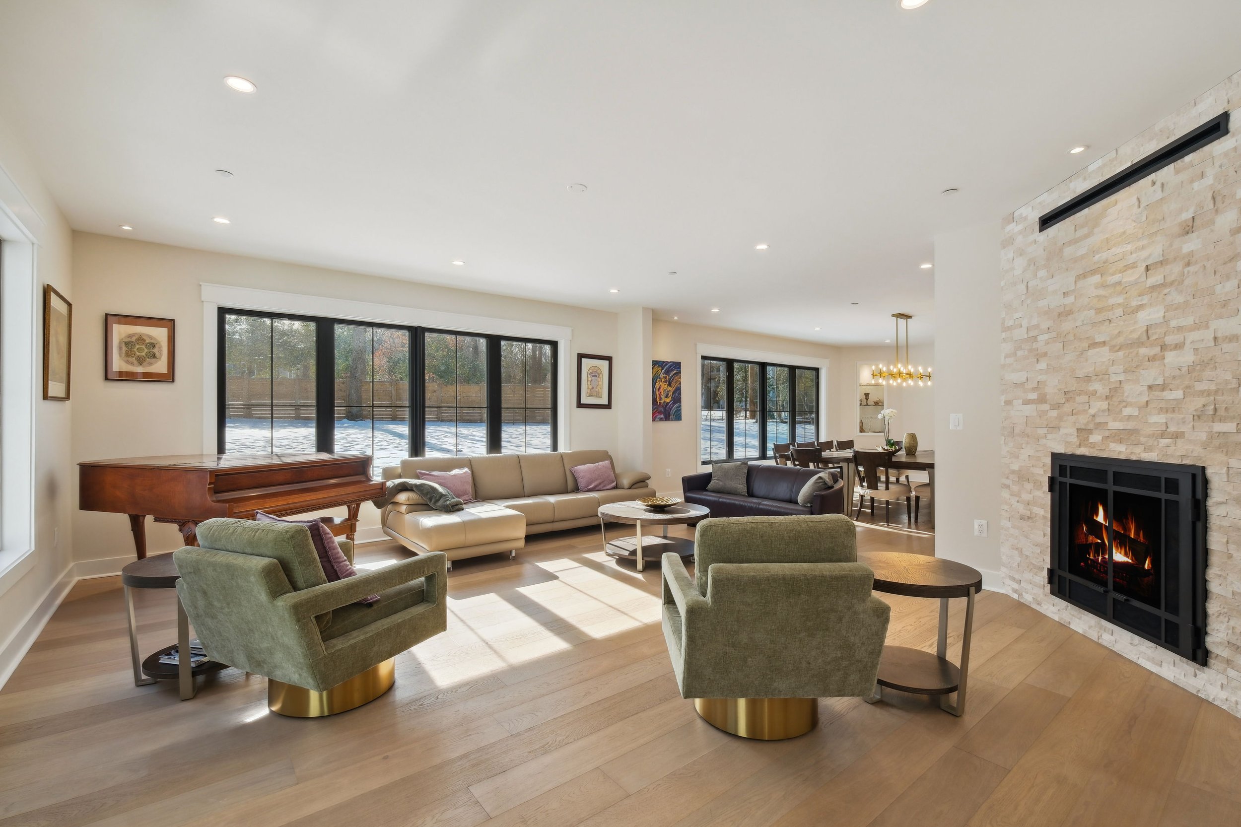 Living room with beige sofas, green armchairs on gold bases, a grand piano, and artwork on the walls. Large windows show an outdoor snowy scene, and a fireplace is built into a beige brick wall. Dining area with a chandelier is visible in the background.