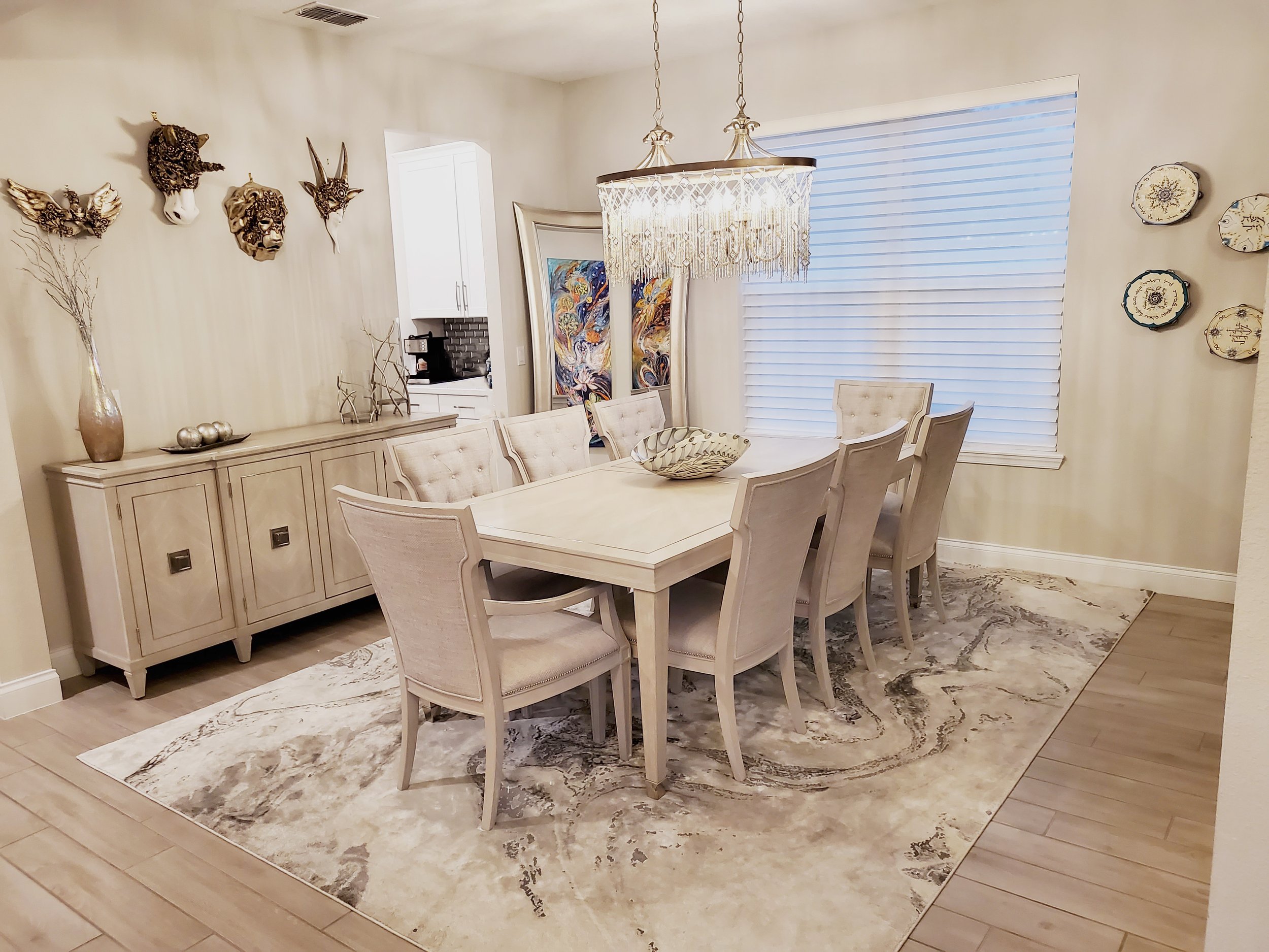 Dining room with a rectangular wooden table surrounded by eight beige upholstered chairs, a light-colored sideboard with decorative items, a patterned area rug, a chandelier, a window with blinds, and wall art including masks and plates.