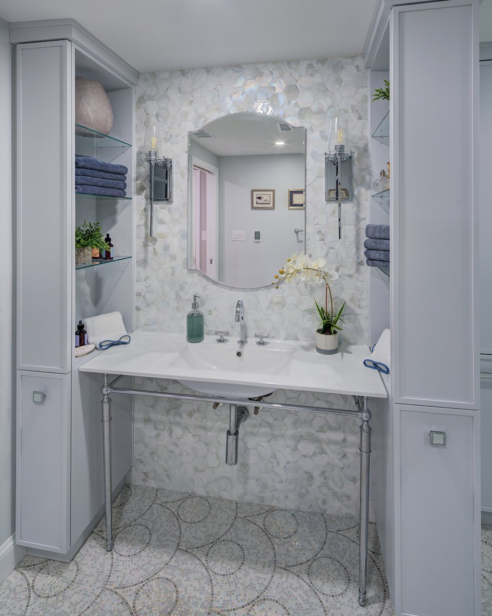Modern bathroom vanity with a white countertop, decorative hexagon tiles on the wall, shelves with towels, plants, and toiletries, and a large mirror.
