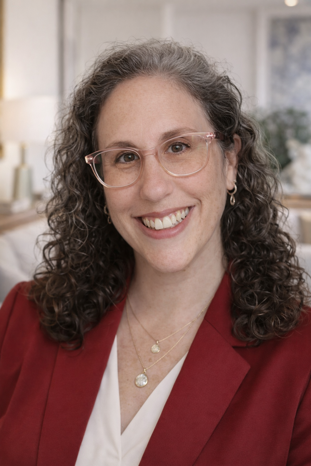 A woman with curly brown hair wearing glasses, gold layered necklaces, earrings, a white top, and a red blazer, smiling in a well-lit indoor setting.