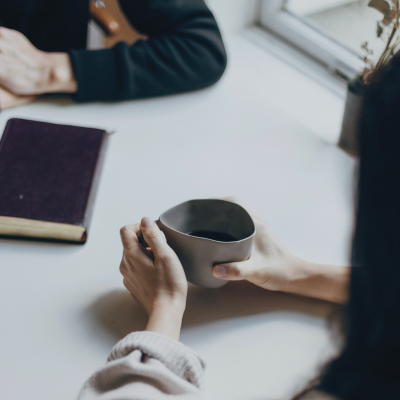 A person holding a dark ceramic mug at a white table, opposite another person with a closed book nearby, in a 1:1 coaching session.