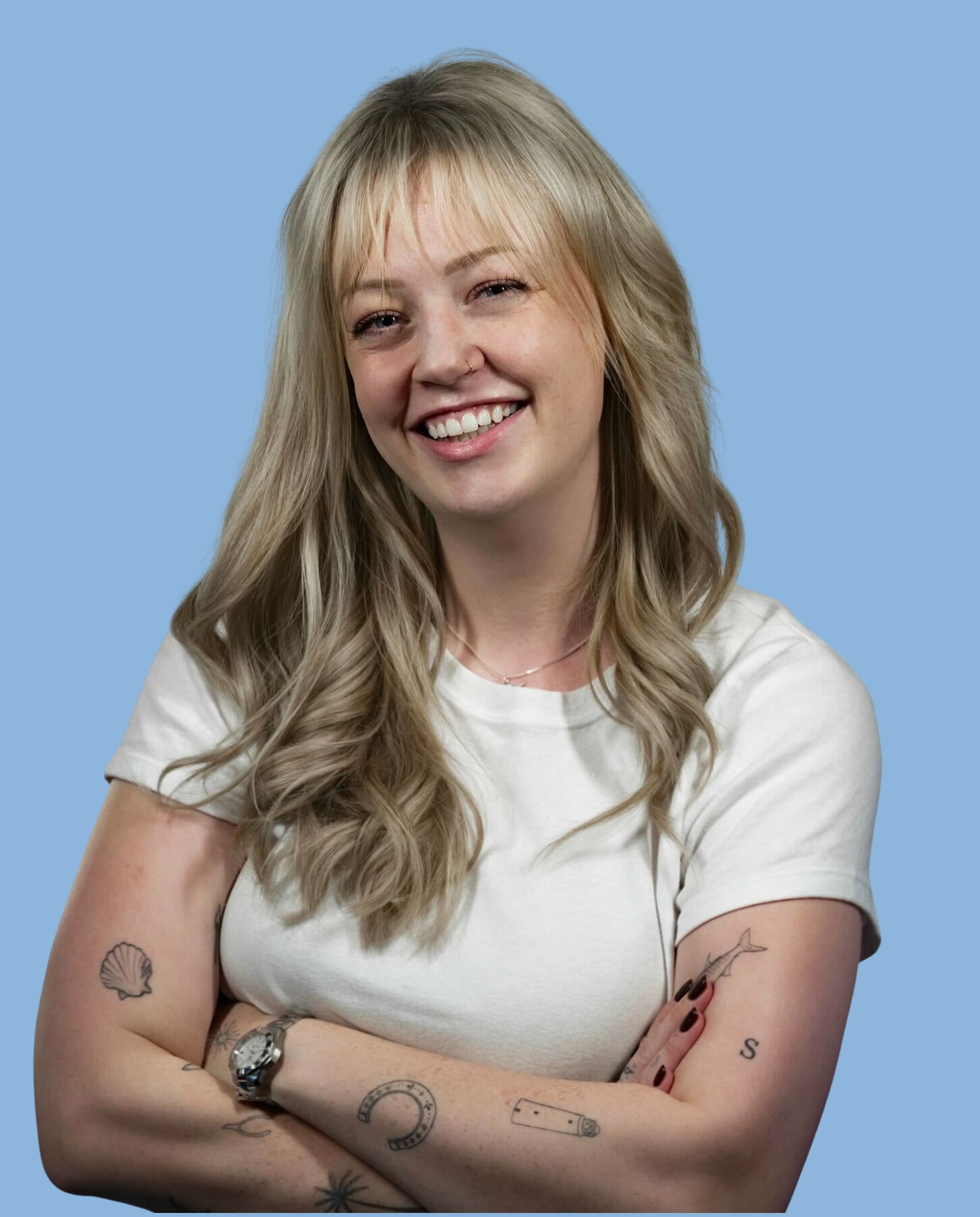 A blonde lady smiling with arms crossed, wearing a white t-shirt against a light blue background.