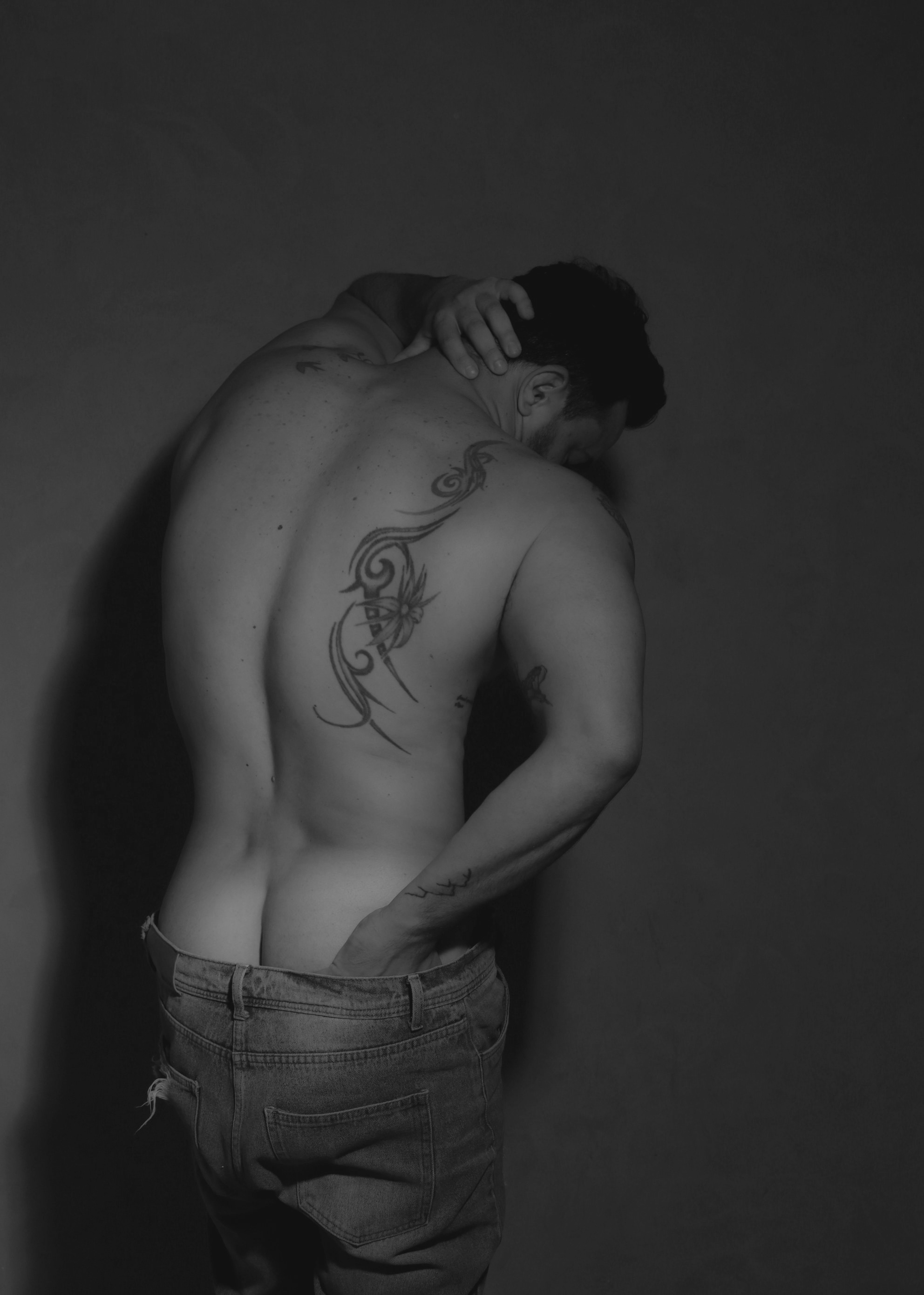 Shirtless man shown from behind in low light, with a large decorative back tattoo, hand resting on his neck, and jeans pulled slightly down to reveal the top of his buttocks.