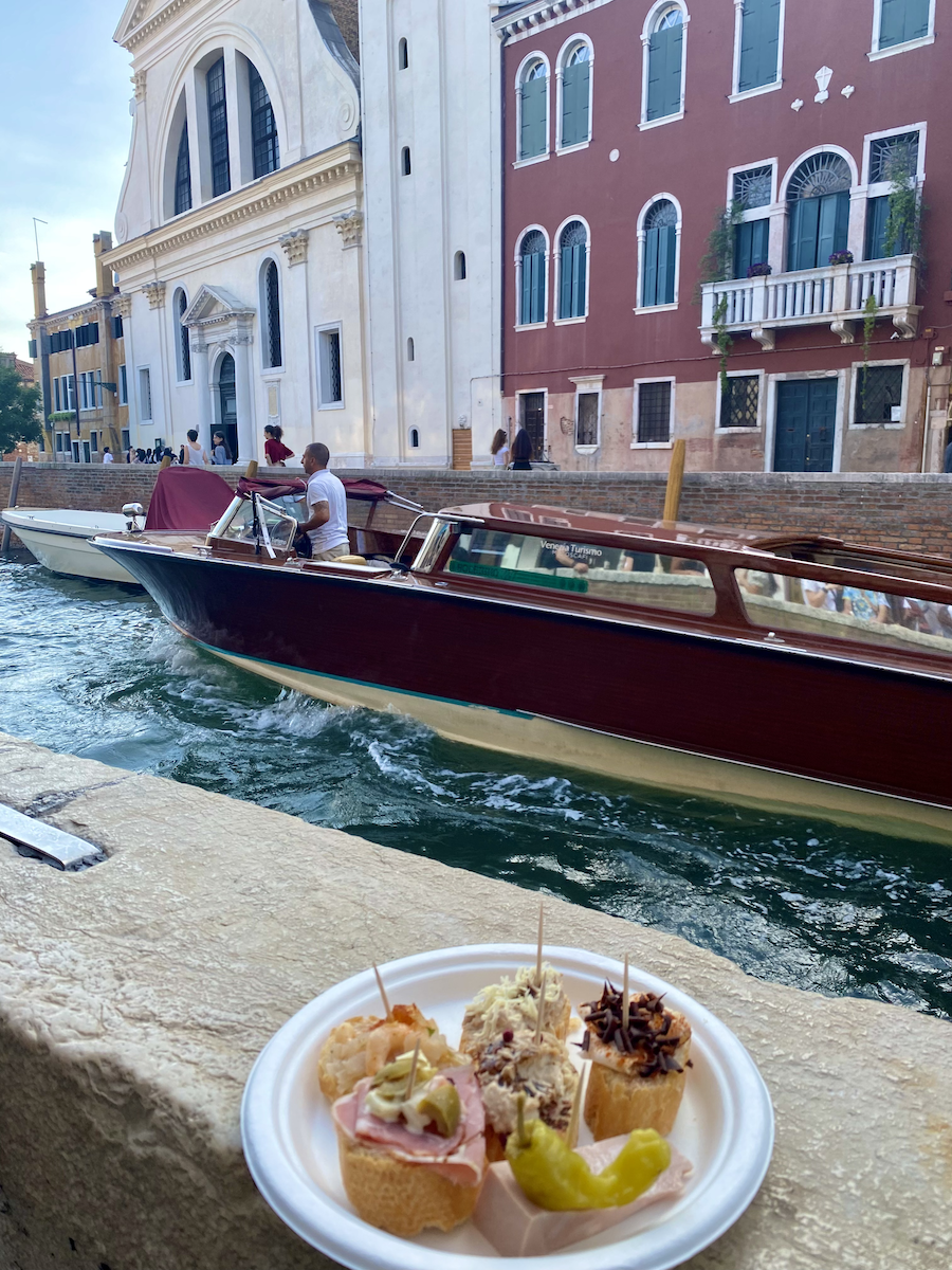 A plate of assorted appetizers with a canal and boats in Venice in the background.