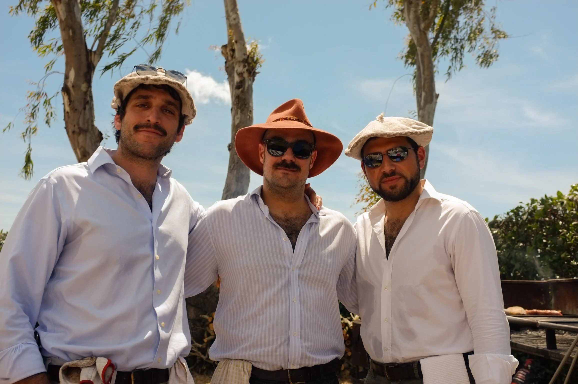 Tre uomini vestiti casualmente con camicie bianche, cappelli e occhiali da sole, che sorridono e si abbracciano all'aperto sotto un cielo blu, con alberi sullo sfondo.