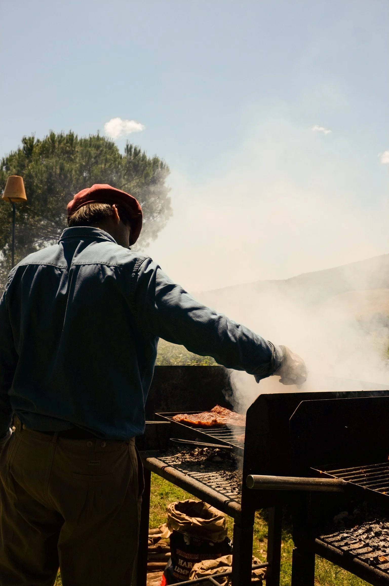 Persona che cucina carne sulla griglia all'aperto con fumo, in un ambiente naturale con alberi e cielo azzurro.