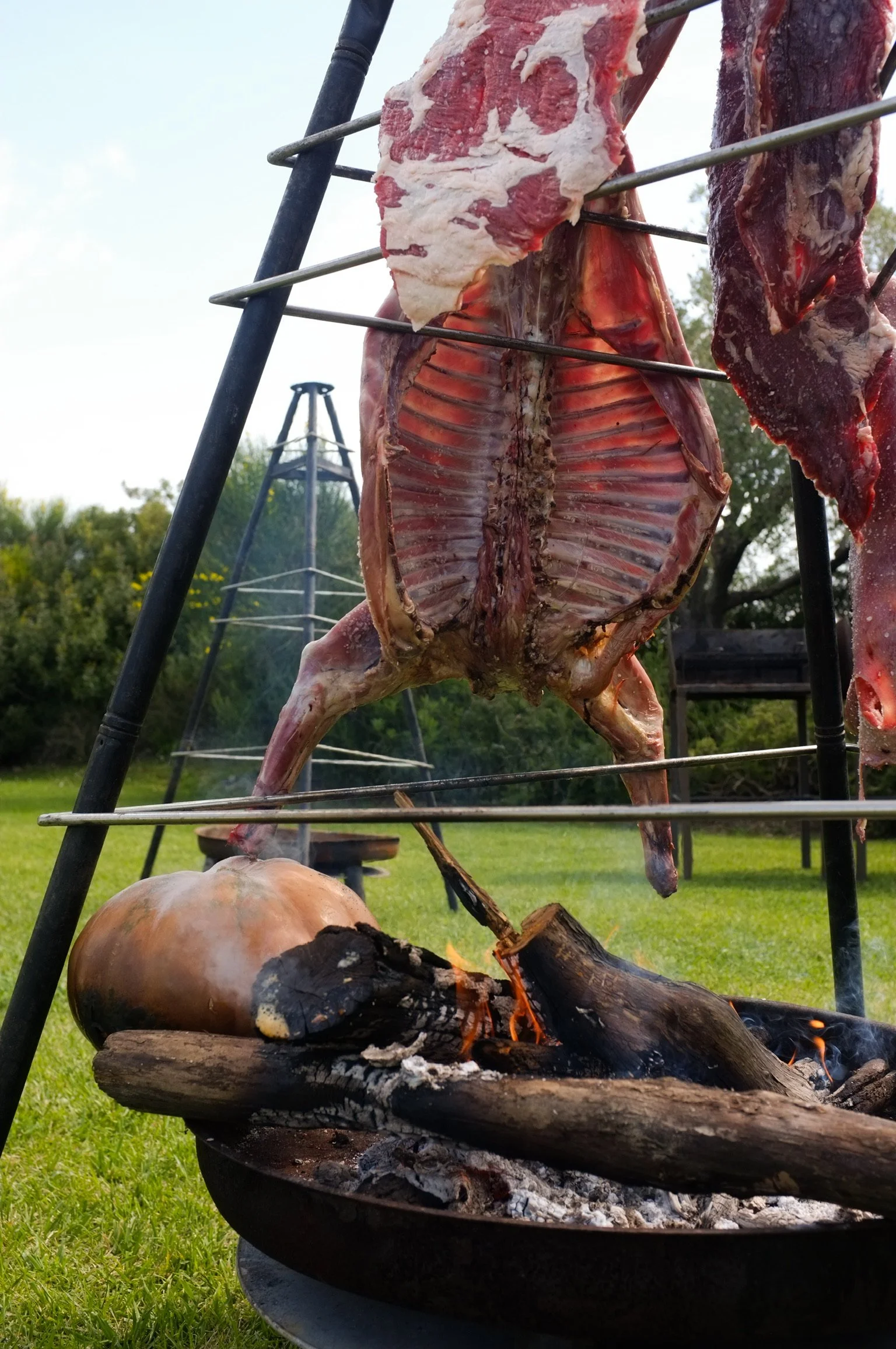 Carne appesa ad un supporto metallico sopra un falò all'aperto.