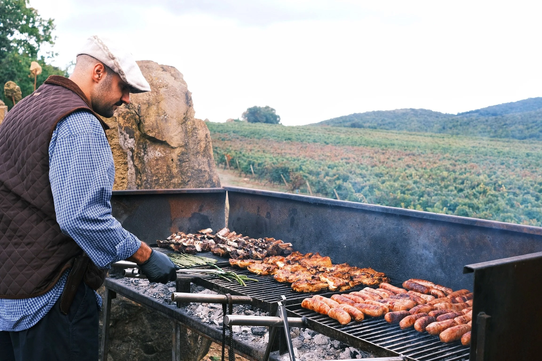 Un uomo sta grigliando carne e salsicce su un barbecue all'aperto con un paesaggio di colline e campi verdi sullo sfondo.
