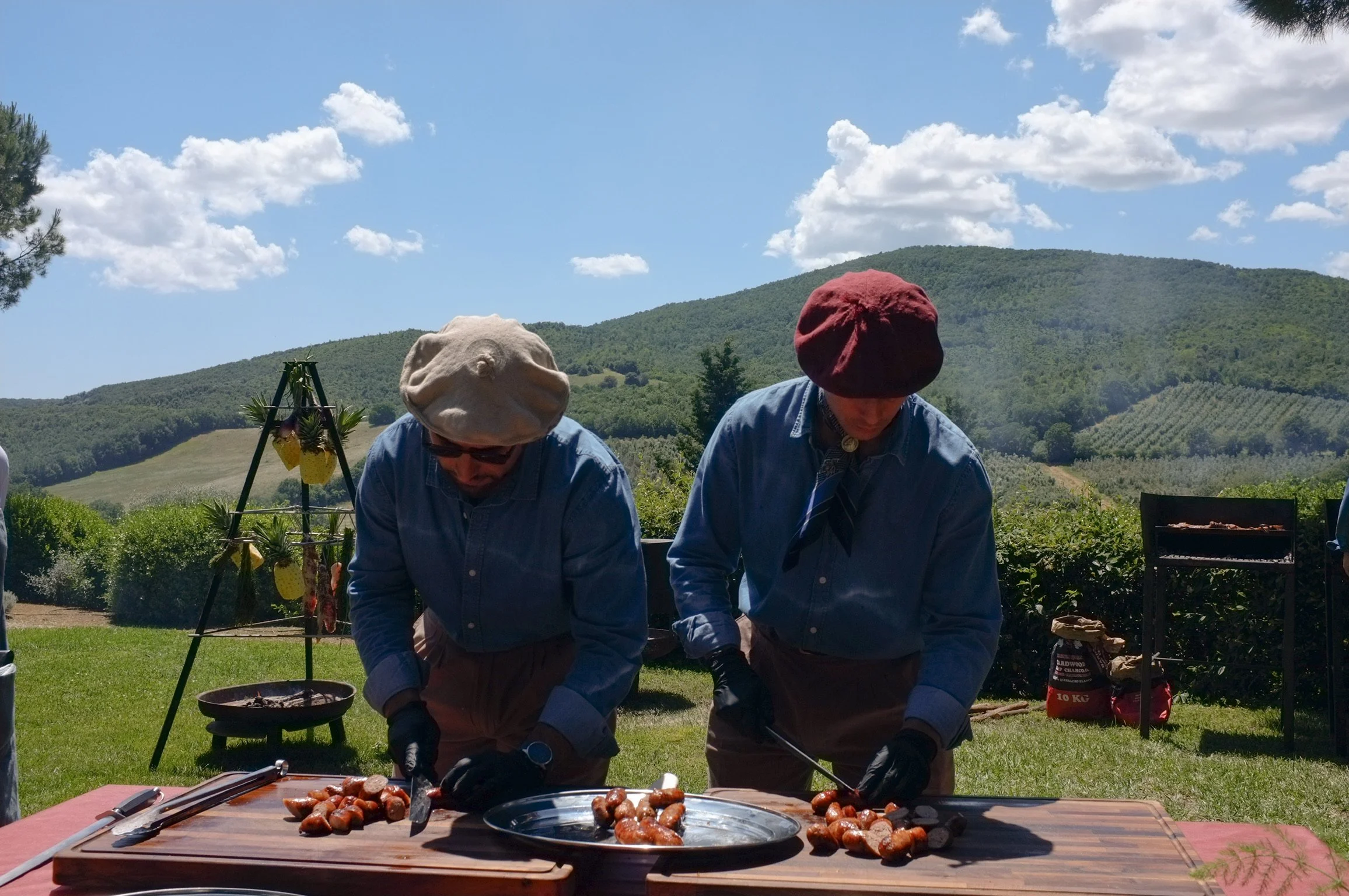 Due persone che preparano e cucinano salsicce su un tavolo di legno all'aperto, con uno sfondo di colline verdi e un cielo blu con nuvole.