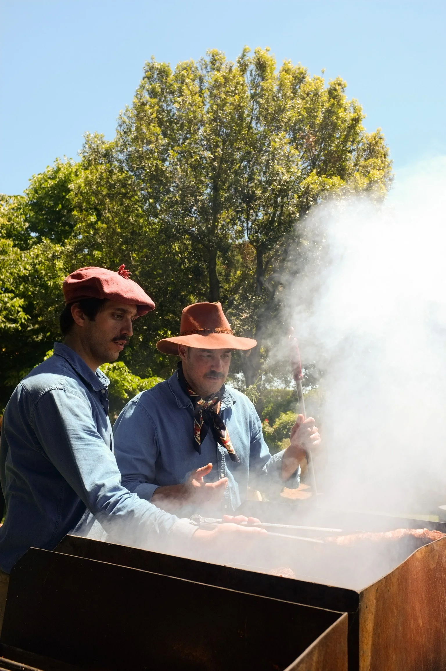 Due uomini vestiti in stile western, con cappelli di paglia, stanno cucinando carne su un barbecue all'aperto. Ci sono alberi e un cielo azzurro sullo sfondo.