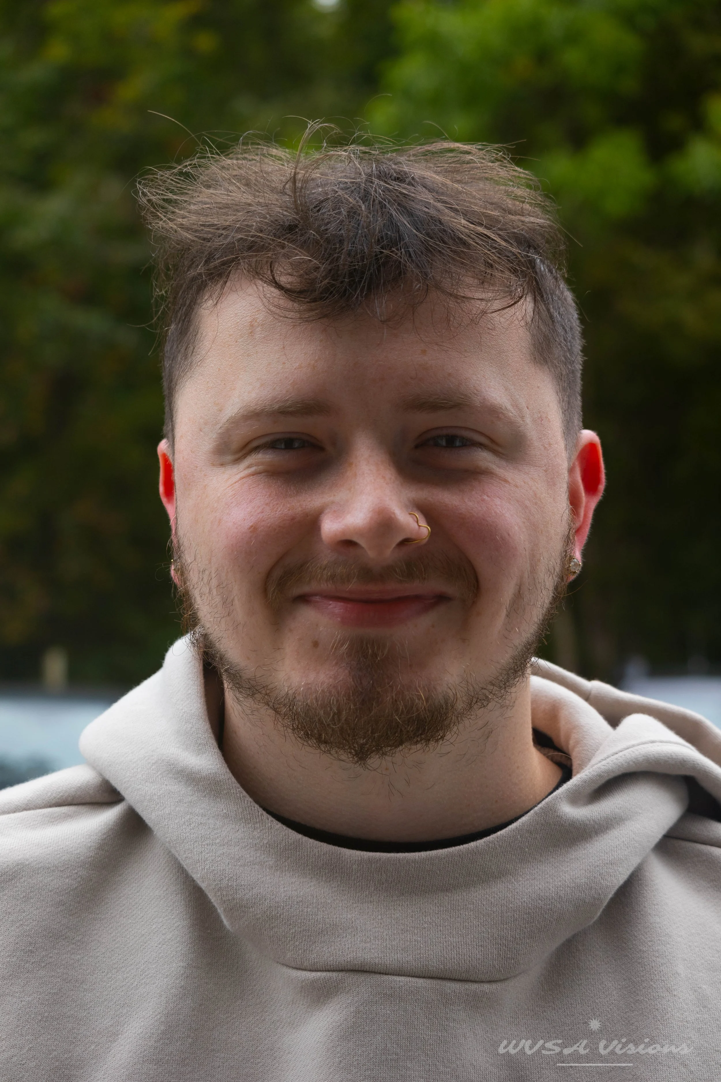 Close-up of a smiling young man with short, curly hair, a nose ring, and earrings, wearing a light beige hoodie outdoors with trees in the background.
