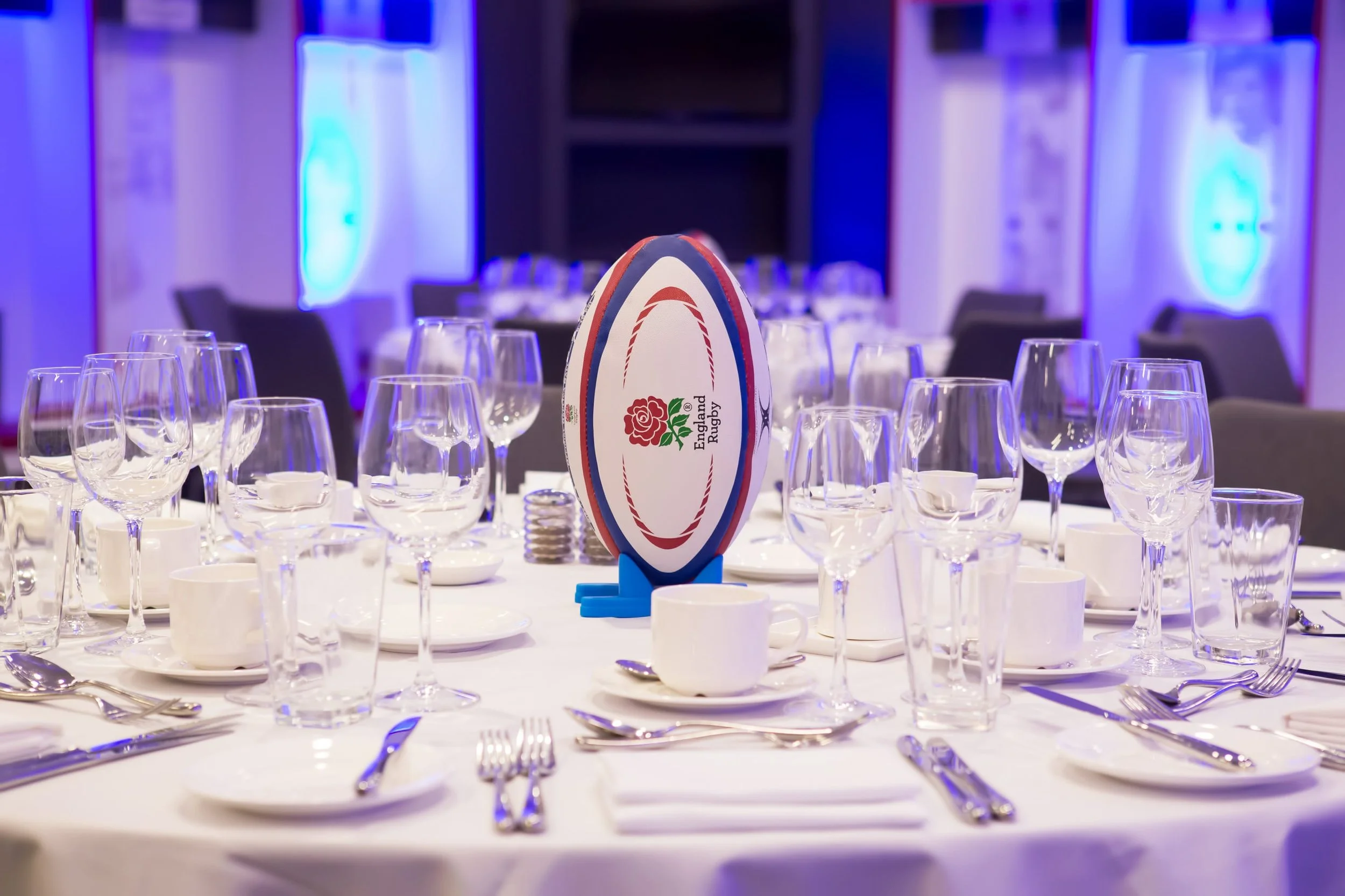 A banquet table set for a formal event with numerous wine glasses, cutlery, and white plates. A rugby ball with the England Rugby logo and a red, white, and blue design stands at the center of the table. The background features a dimly lit room with blue lighting and large, round logos on the walls.