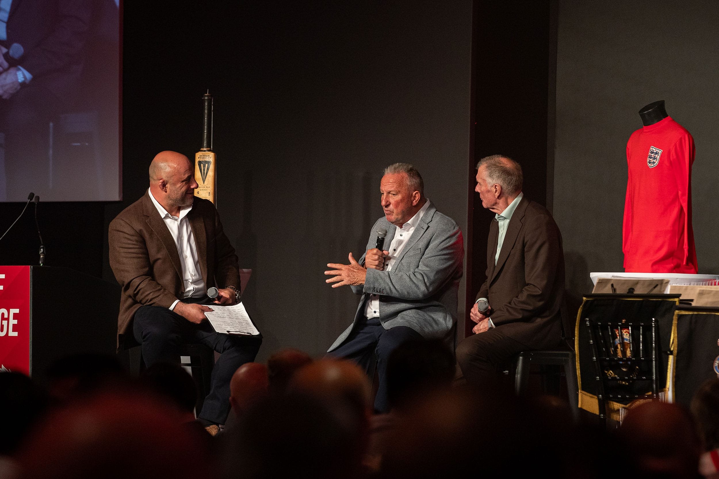Three men seated on stage during a panel discussion, with a fourth man standing and speaking into a microphone. The stage features a display of a red England football jersey and a football-related item.