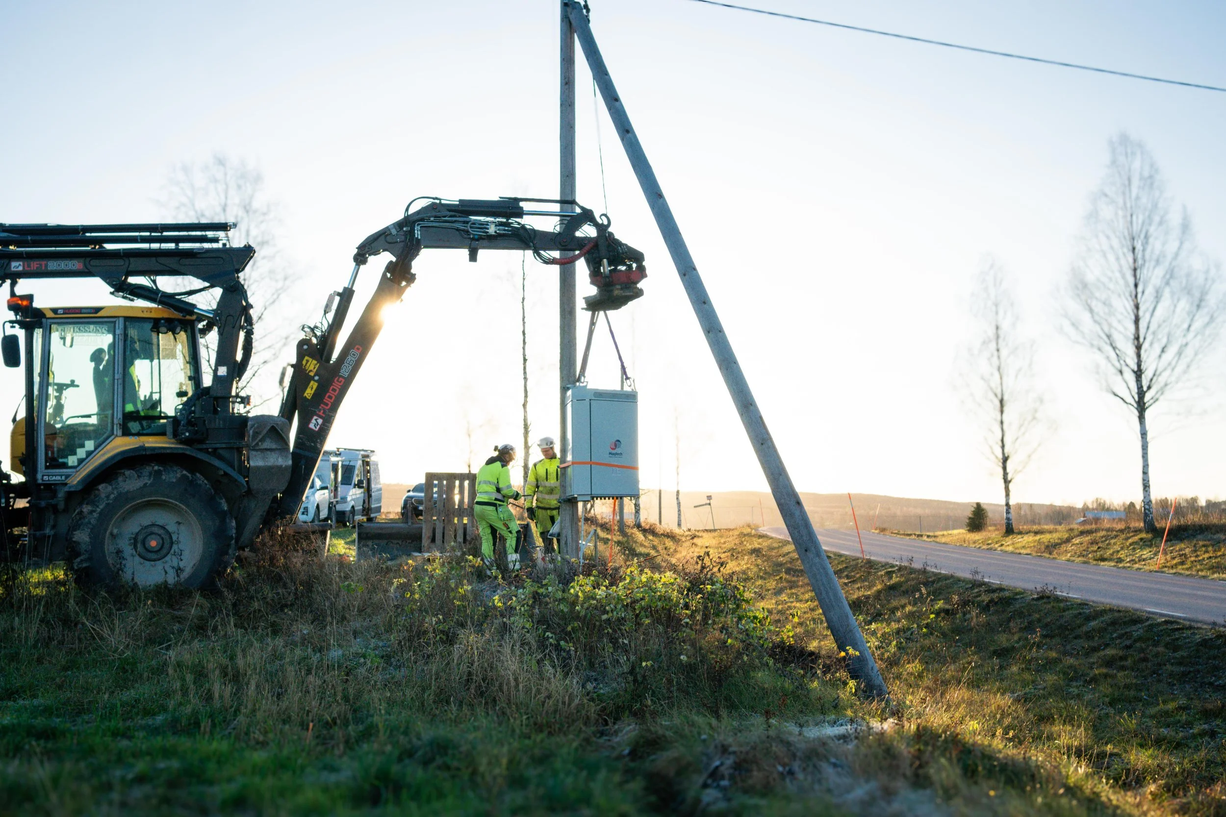 En traktor som håller i en spänningsreglerande enhet och två montörer vid en stolpe.