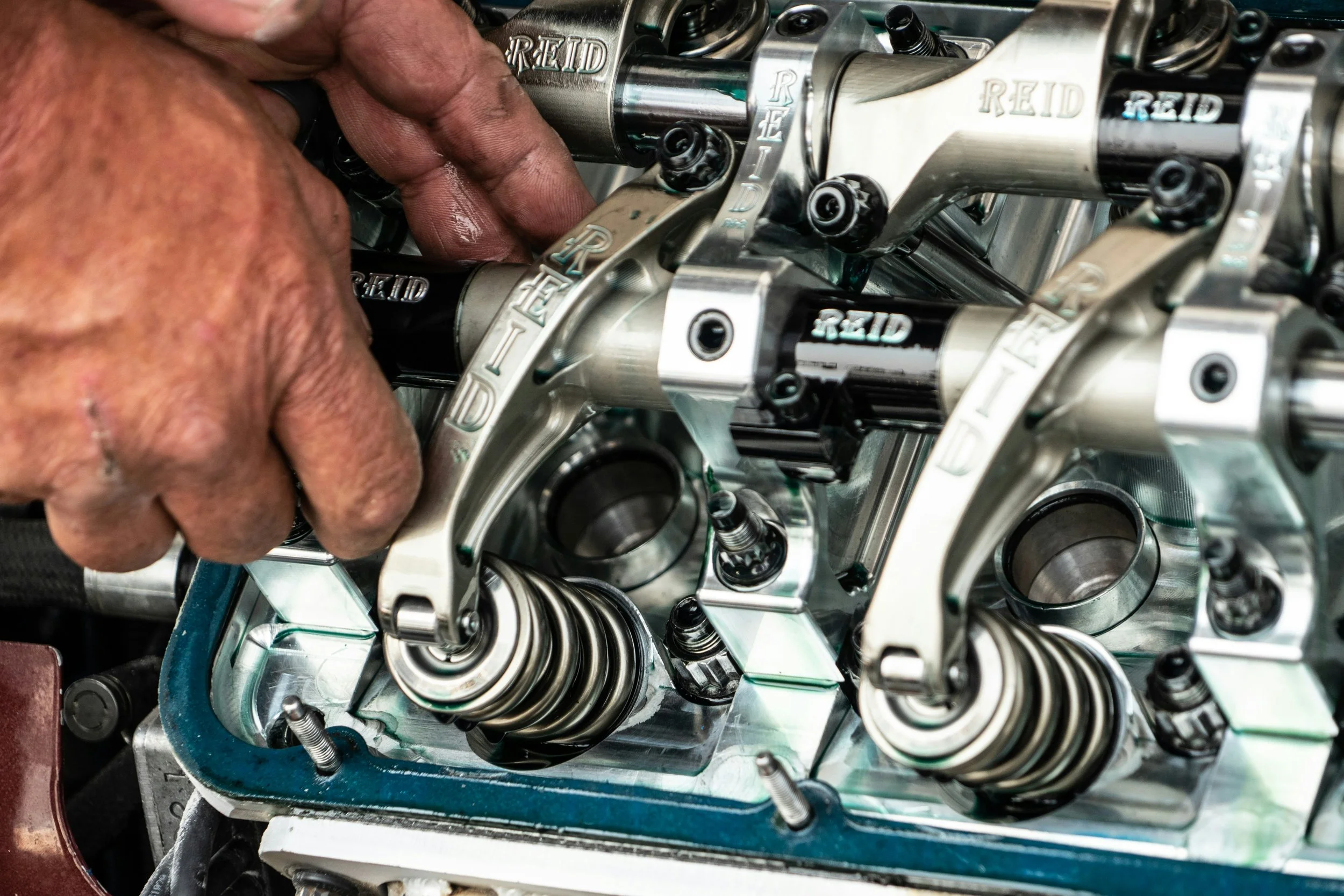 Close-up of a mechanic's hand working on a high-performance engine's valve and rocker arm assembly, with visible engine components and branded parts.