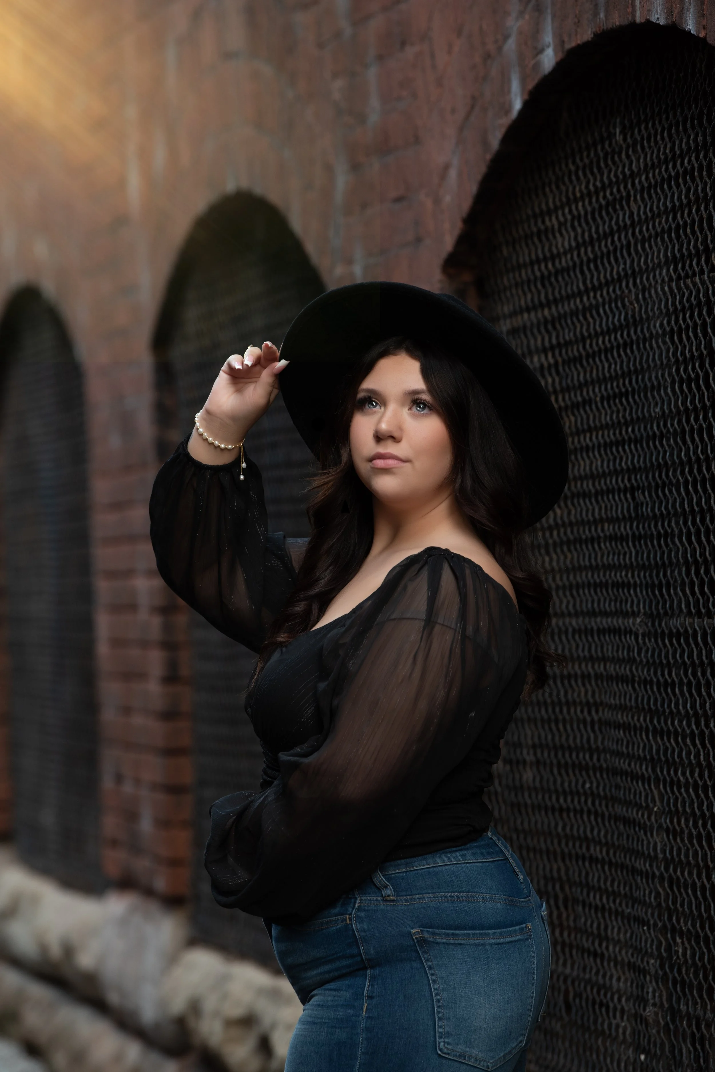A young woman with long dark hair, wearing a black sheer blouse, jeans, and a wide-brimmed black hat, poses against a brick wall with black mesh window coverings, looking off into the distance.