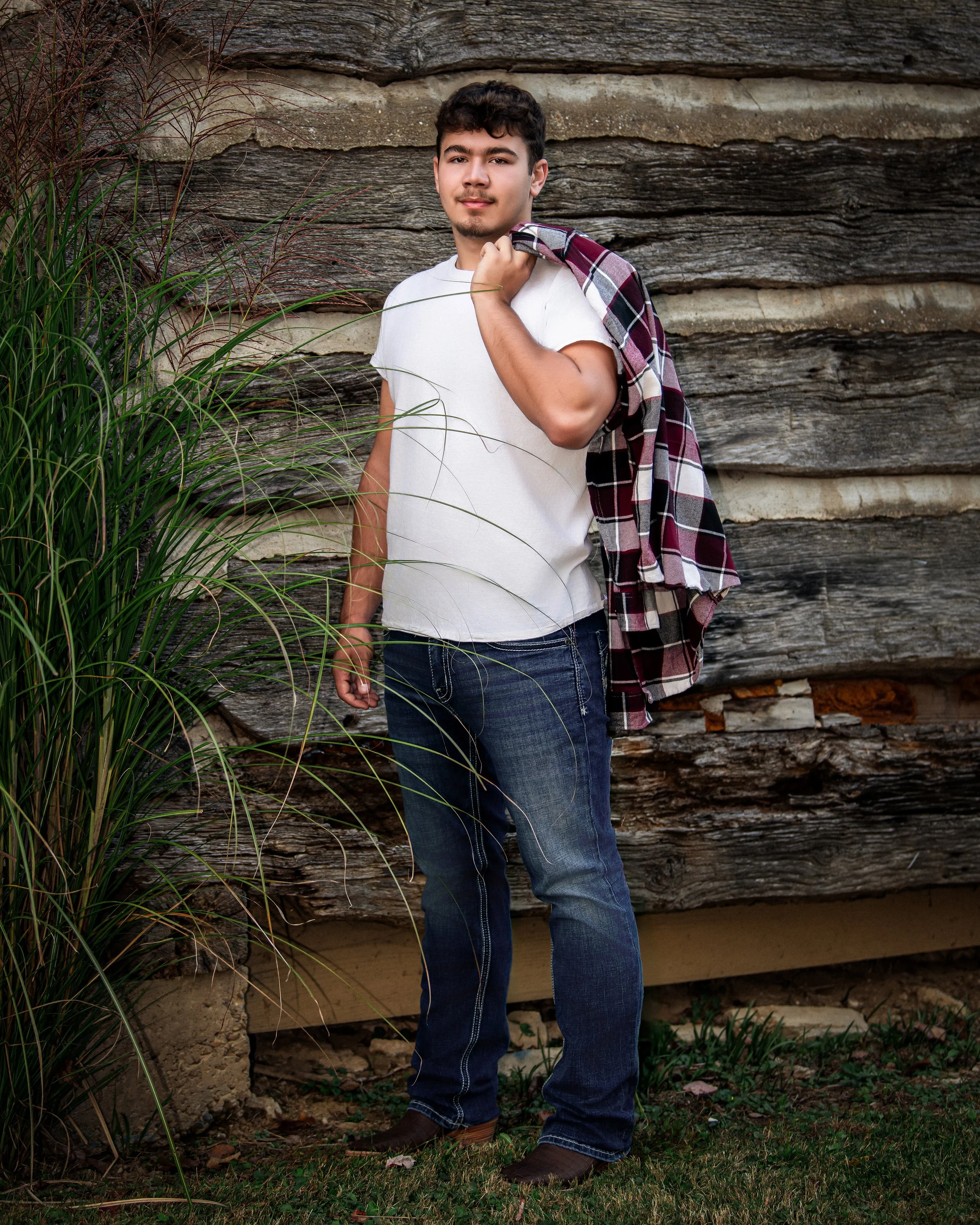 A young man with dark hair and a mustache stands outdoors in front of a weathered wooden wall, holding a plaid shirt over his shoulder, wearing a white t-shirt and blue jeans.