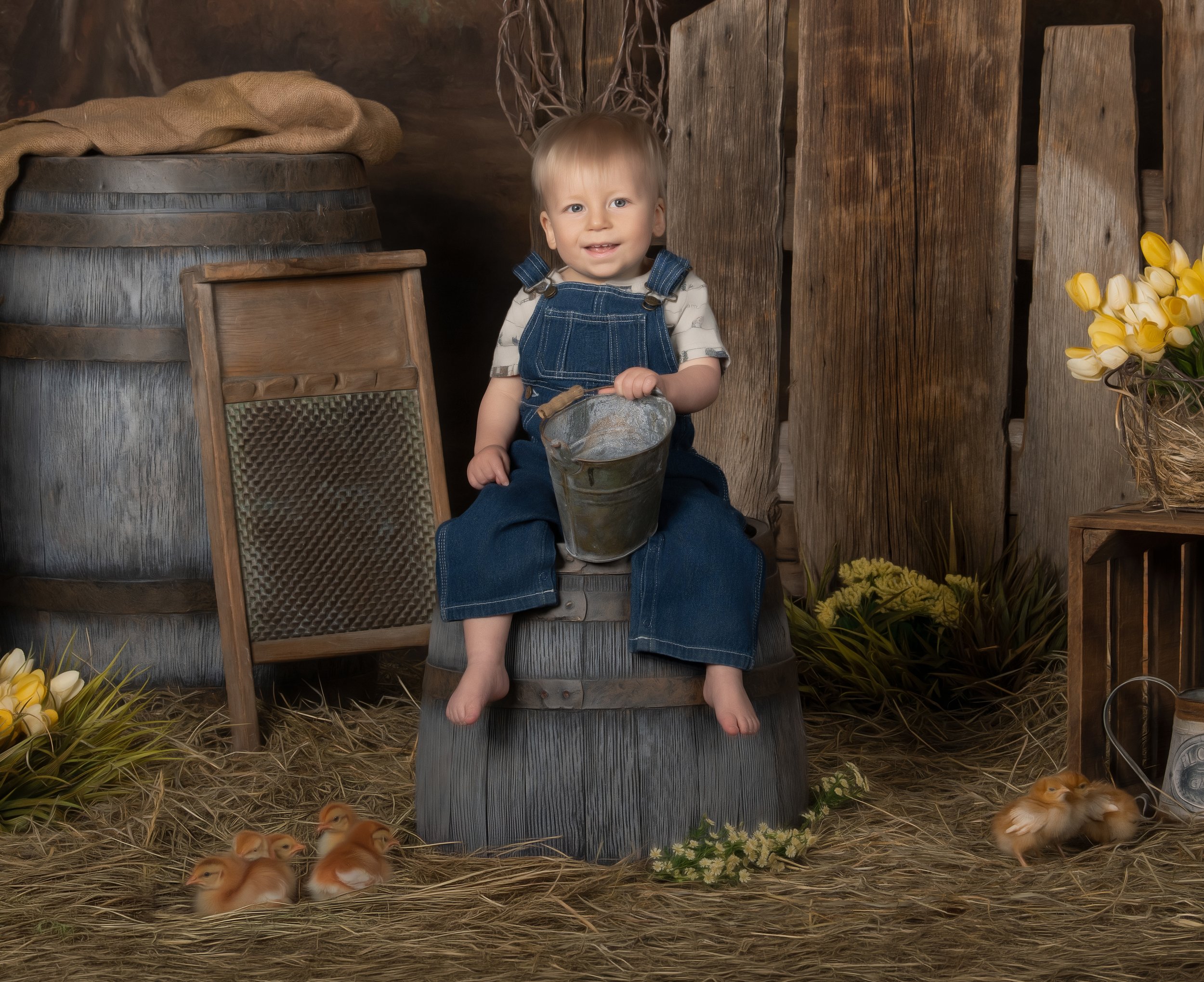 A young child sitting on a wooden barrel in a rustic farm setting, holding a metal bucket, with ducklings on the straw-covered floor around them, wooden crates, flowers, and farm-related items in the background.