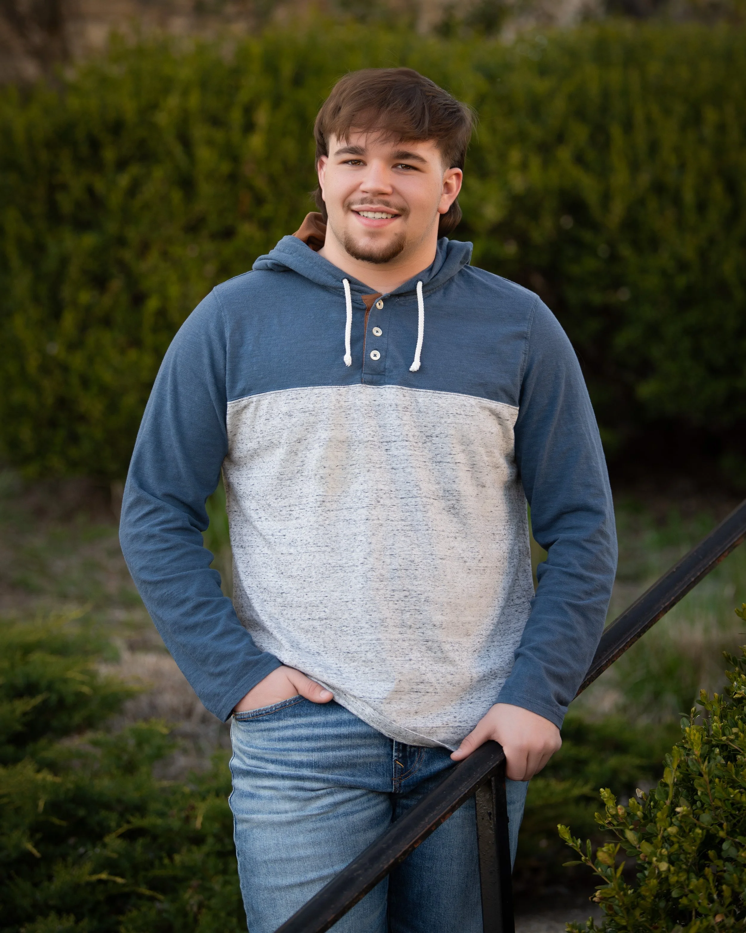 A young man in a blue and gray hoodie and jeans smiling outdoors with bushes and trees in the background.