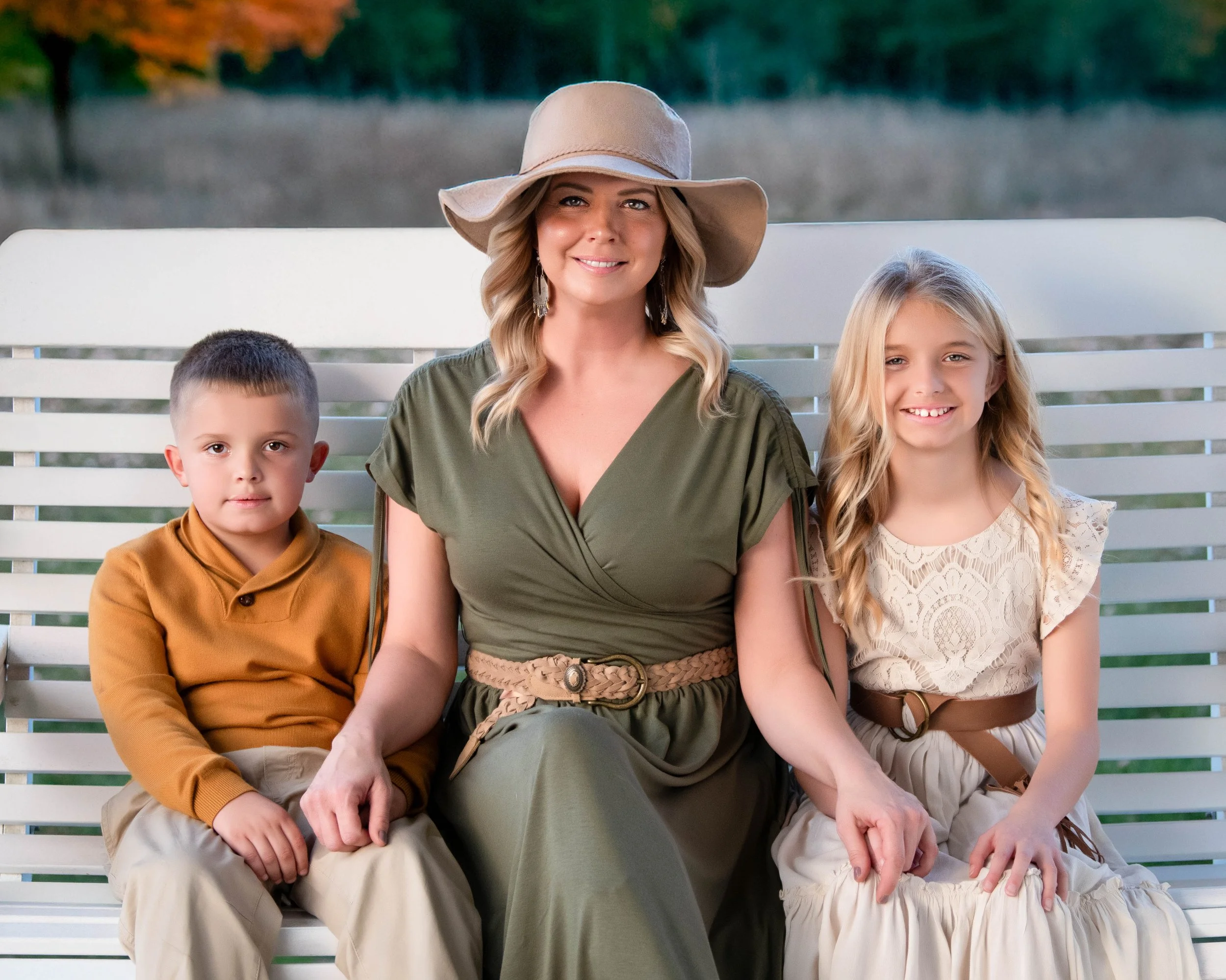 A woman with blonde hair wearing a wide-brimmed hat and green dress, sitting on a white bench with a young boy in an orange shirt and beige pants on her left, and a young girl in a white lace dress and beige skirt on her right.
