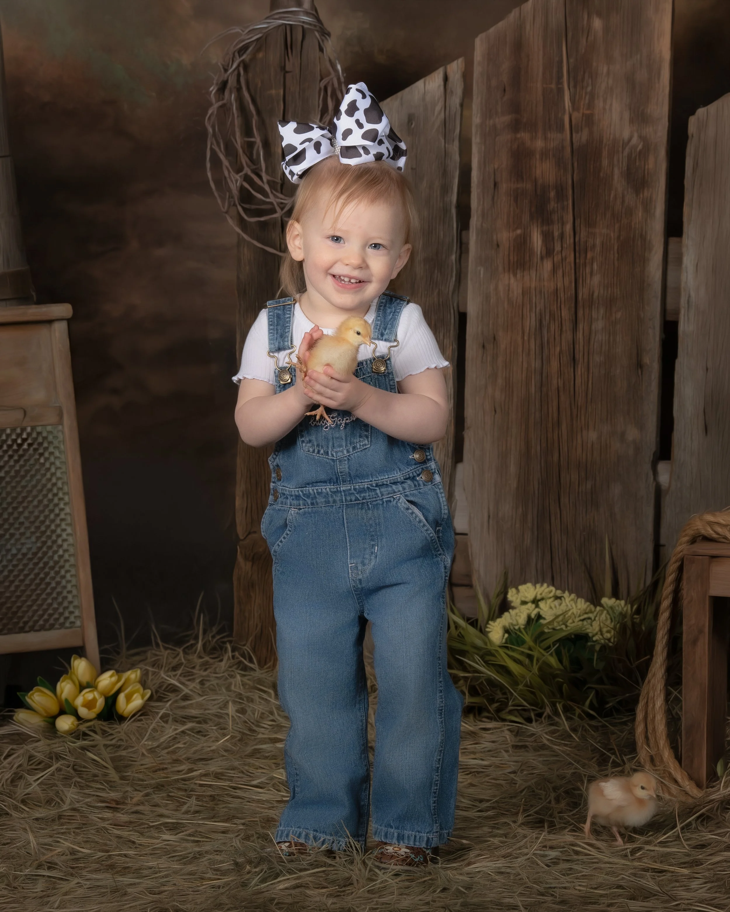 A young girl with a big polka-dot bow in her hair, wearing denim overalls and a white shirt, is holding a yellow chick and smiling. She stands in a rustic farm setting with hay on the ground, wooden fence, and a small chick on the floor.