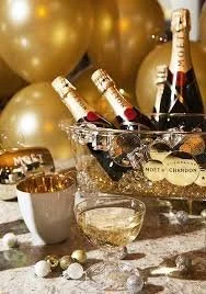 Chilled bottles of champagne in a silver bucket, gold balloons in the background, and a tray with glasses and small candies on a decorated table, suggesting a celebration or party.