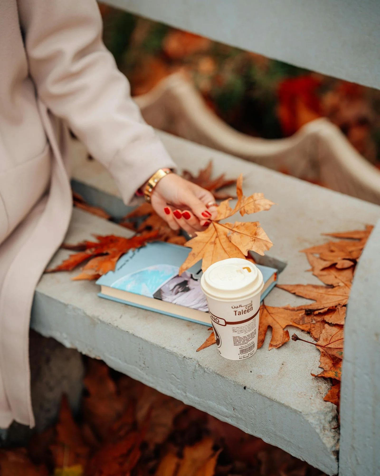 A woman sitting on a bench holding autumn leaves beside a book and a takeaway coffee, creating a quiet, reflective moment.