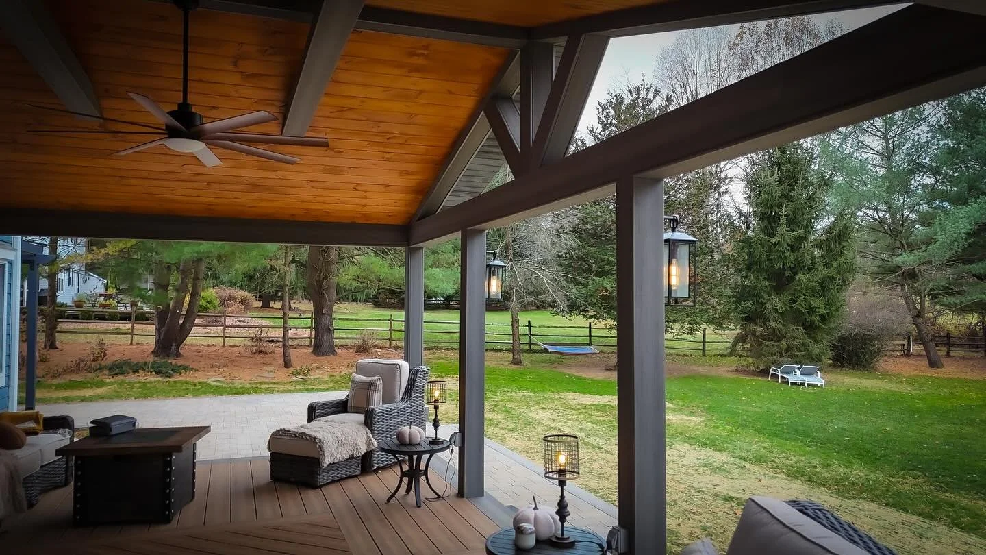 Covered backyard porch with seating area, ceiling fan, lanterns, and a view of a fenced grassy yard with trees and outdoor furniture.