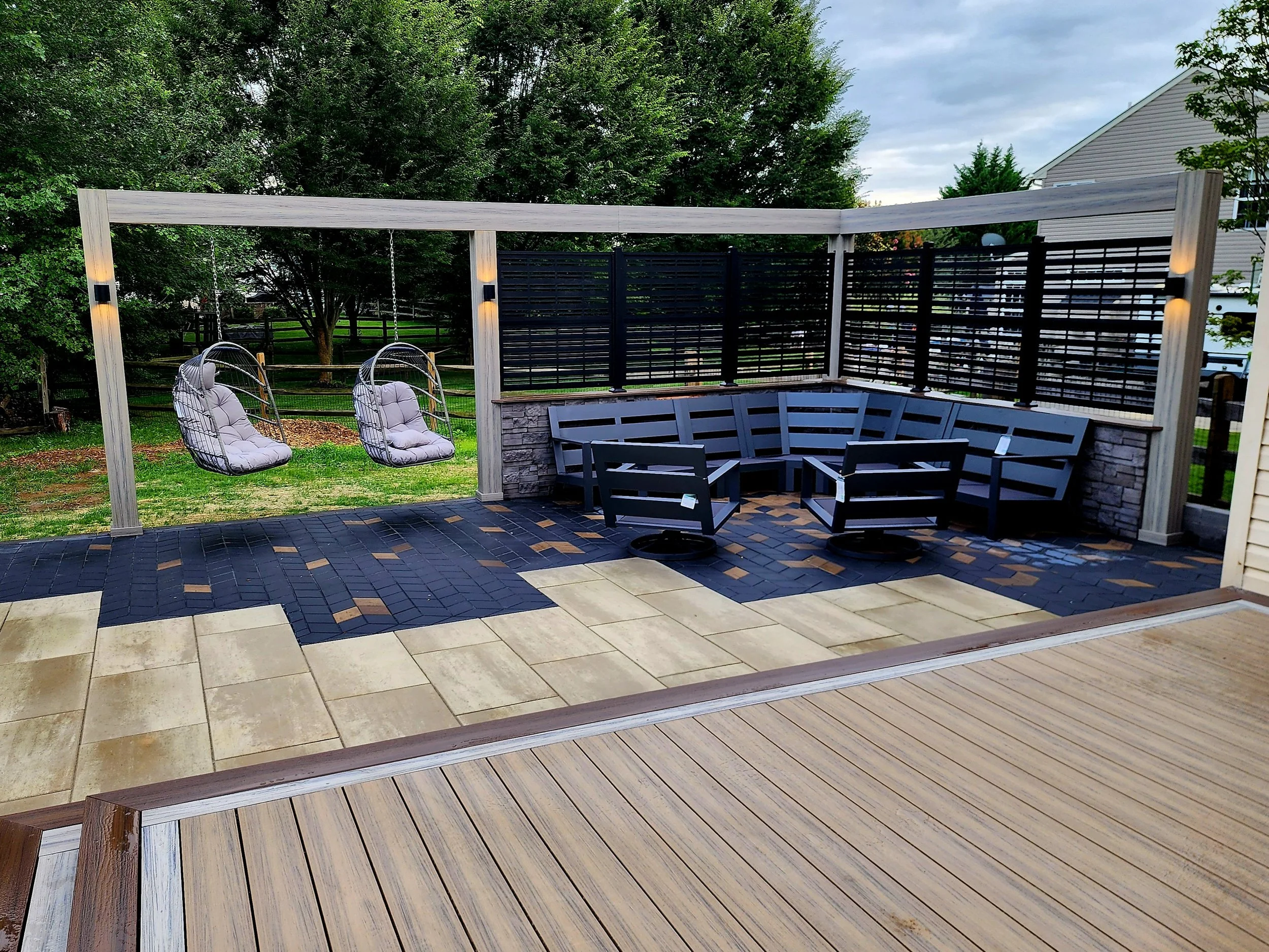 Backyard patio featuring two hanging egg chairs with cushions, a curved black fence, a bench with armrests, and a wooden deck area with pavers and grass in the background.