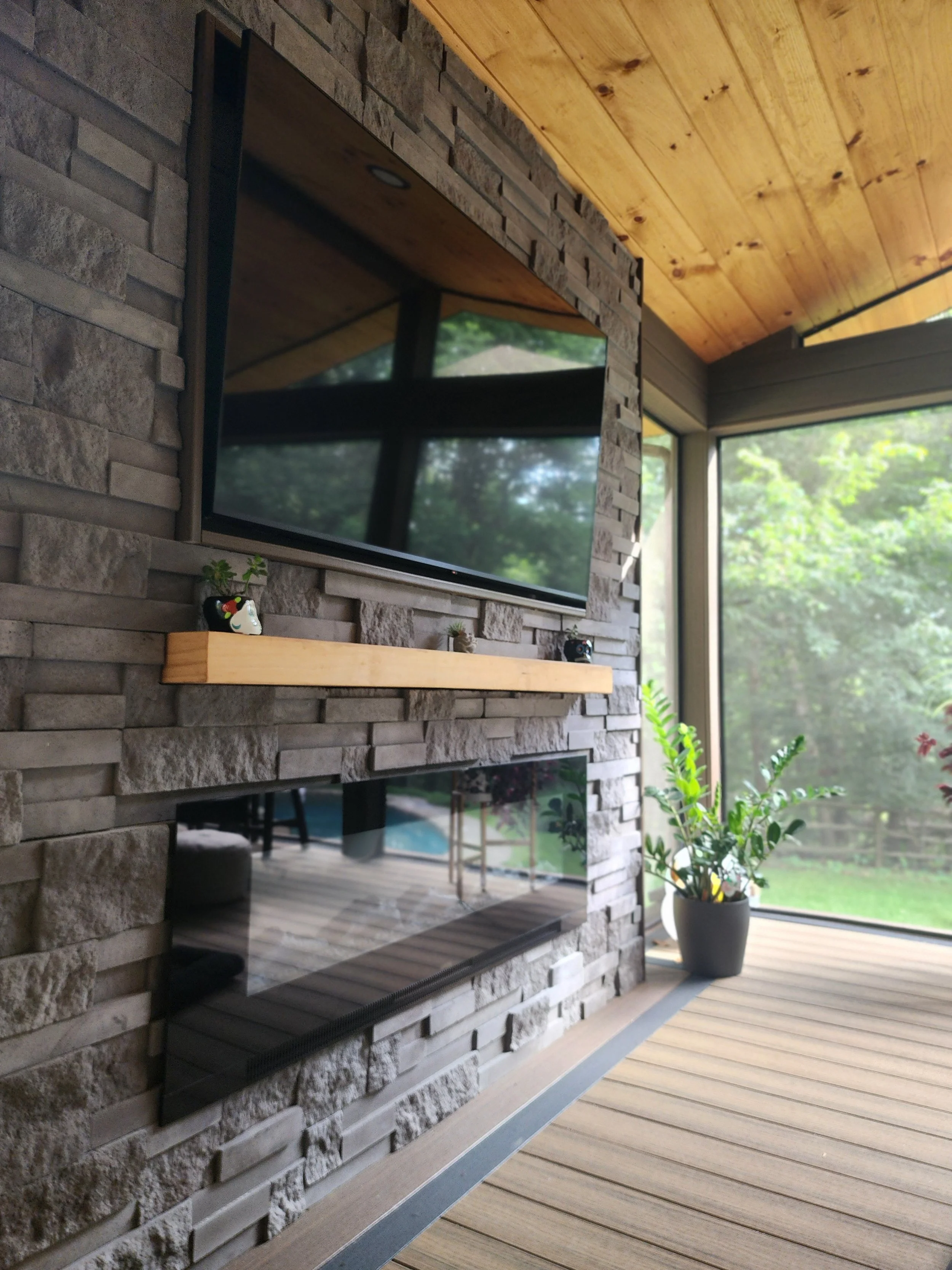 Living room with a wall-mounted flat-screen TV above a modern fireplace with a stone surround and wooden mantel, next to large windows with a view of green trees, and a potted plant on the wooden floor.